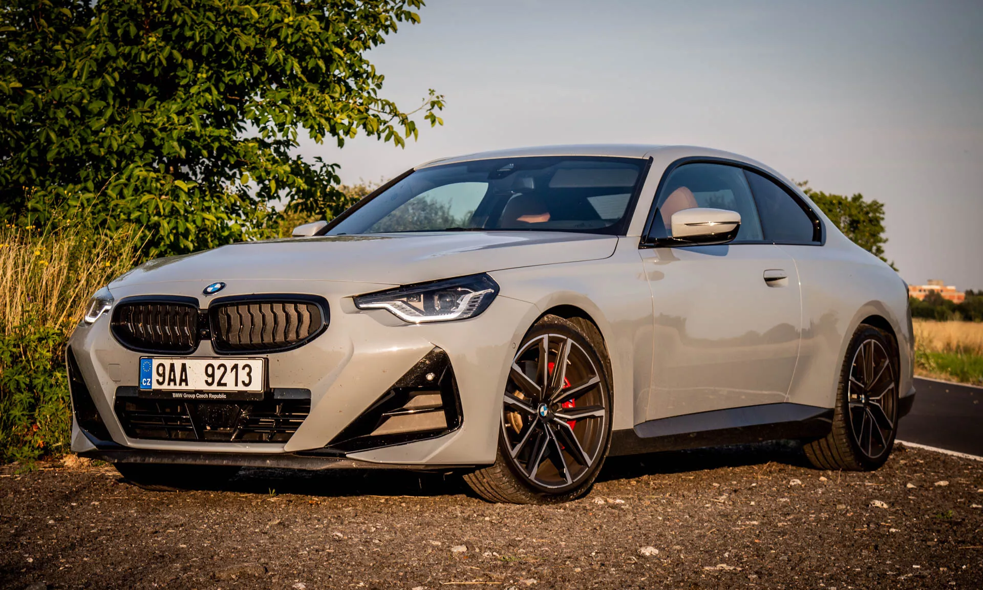 Image d'une BMW Série 2 Coupé 2022. La voiture de luxe moderne est garée en angle, mettant en valeur le côté avant-gauche au cours de l'heure dorée. L'image se concentre sur des éléments de design distinctifs comme la grande calandre en forme de rein, les phares LED angulaires, le pare-chocs avant sportif avec de grandes prises d'air et les roues en alliage noir à plusieurs rayons avec des étriers de frein haute performance. Le rétroviseur aérodynamique intègre des clignotants. La carrosserie élégante et la finition de peinture premium soulignent le profil compact et sportif de la voiture, ainsi que la ligne de toit basse.