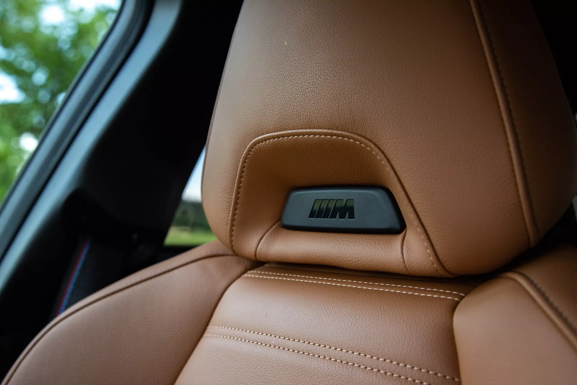 Vue rapprochée de la section supérieure d'un siège en cuir marron riche dans une BMW Série 2 Coupé 2022, mettant en avant les coutures visibles précises et une ligne de contour ergonomique pour le confort et le soutien. Un badge intégré dans un creux sur la partie supérieure du siège indique des fonctionnalités technologiques telles que des réglages électroniques ou des fonctions de chauffage. La prise de vue est réalisée d'un point de vue légèrement inférieur, accentuant la texture luxueuse du cuir tout en floutant l'arrière-plan, qui comprend une partie de la fenêtre de la voiture.