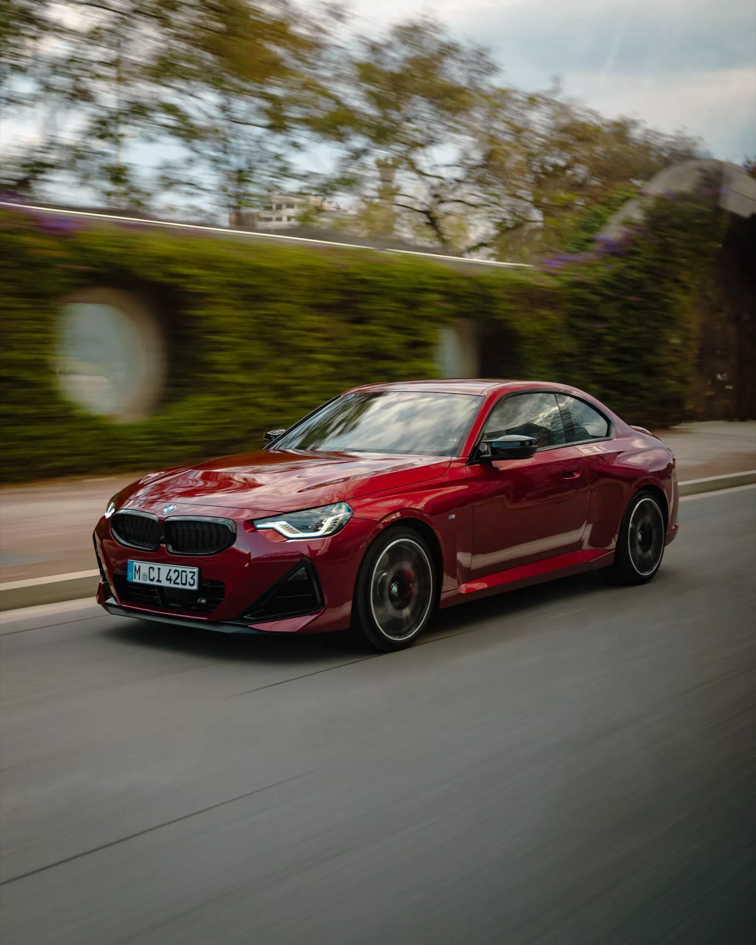 BMW 2 Series 2024 (G42-G87 facelift) rouge vif en mouvement, capturée de profil latéral avec un léger angle arrière. La photo utilise la technique du filé pour le flou de l'arrière-plan, mettant en valeur le design sportif de la voiture, des lignes épurées, des jantes en alliage à rayons multiples, des rétroviseurs de la couleur de la carrosserie et des phares à LED anguleux et acérés. Suggestion d'environnement urbain ou suburbain.