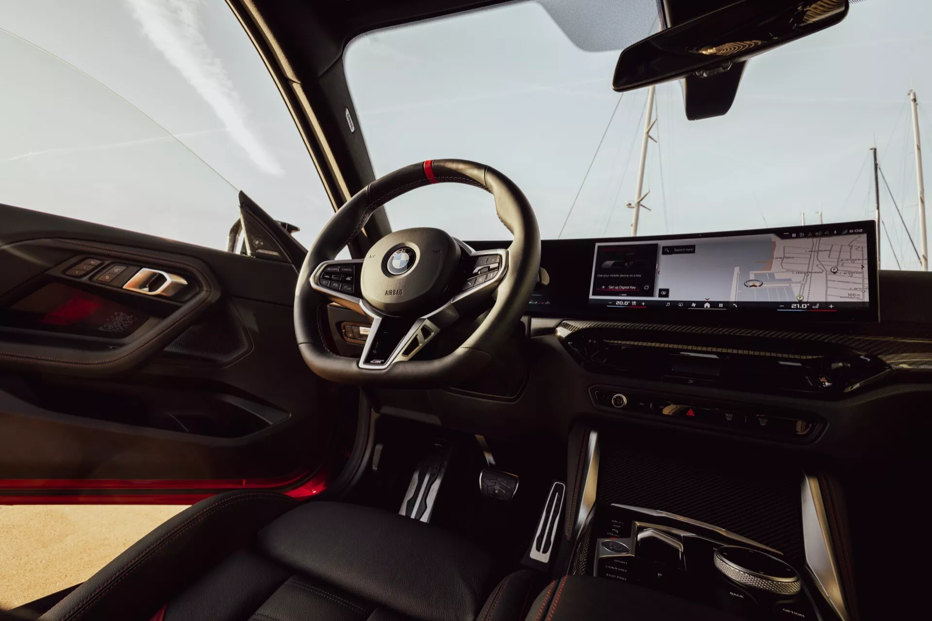 Vue intérieure de la BMW 2er-reihe 2024 (G42-G87 Facelift) depuis le siège passager. L'image montre un volant à trois branches avec des commandes intégrées, un tableau de bord numérique, un grand écran tactile d'infodivertissement et des commandes de climatisation minimalistes. Une illumination d'ambiance rouge est visible et le sélecteur de transmission automatique est situé dans la console centrale. Le panneau de porte comprend des commandes de fenêtres et de rétroviseurs ainsi qu'une grille de haut-parleur. Les sièges en cuir avec coutures contrastées ajoutent une touche de luxe.