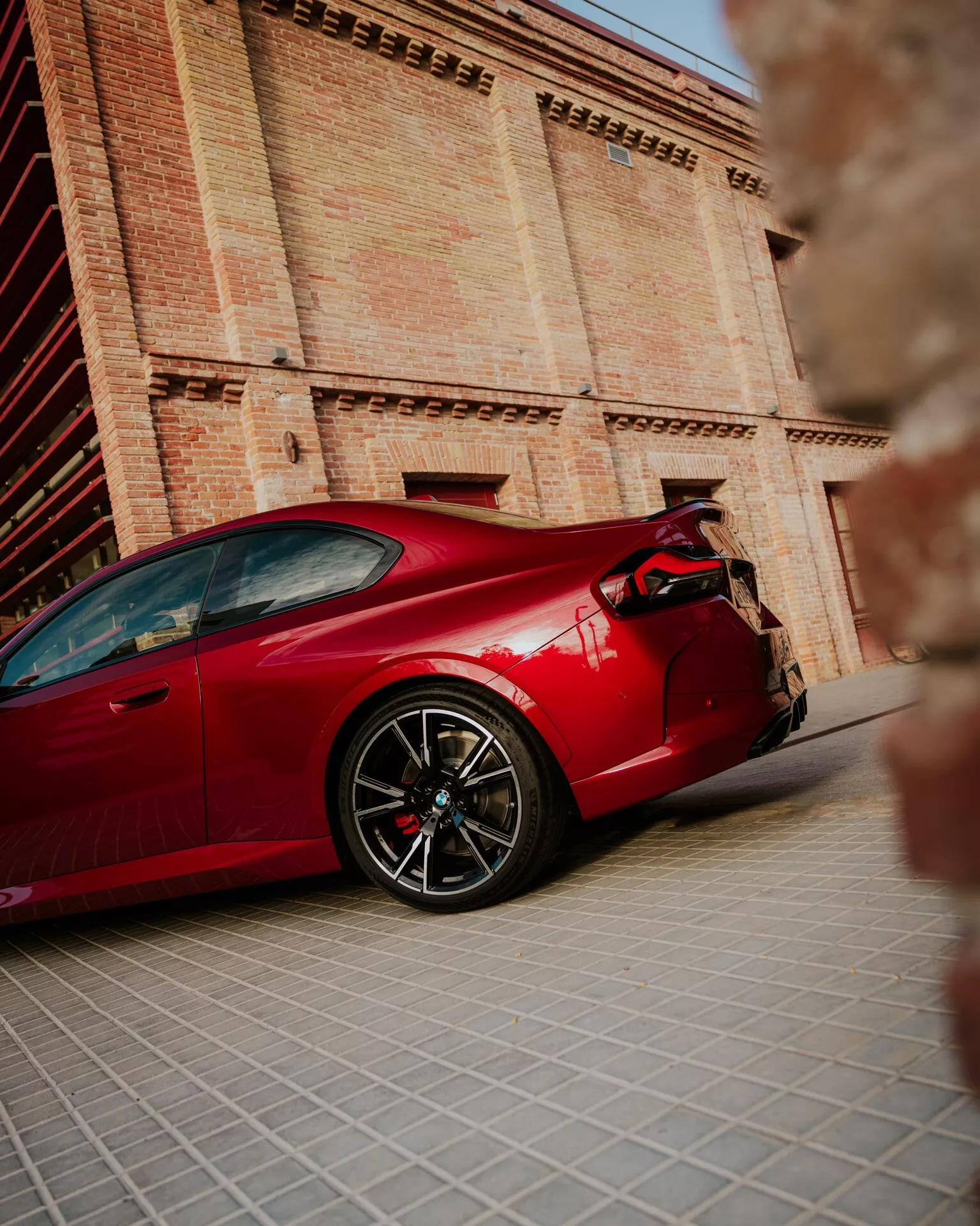 BMW Série 2 2024 (facelift G42-G87) en rouge, capturée sous un angle latéral bas, montrant le profil complet de la voiture, y compris la roue avant, la porte, la prise d'air latérale et la roue arrière. L'image met en évidence les roues haute performance, les rétroviseurs latéraux aérodynamiques, le style de carrosserie coupé élégant, les grandes étriers de frein et le spoiler arrière. Photographie avec une faible profondeur de champ, l'arrière-plan flou présente un bâtiment historique, mettant en avant la voiture comme le point focal principal et soulignant les lignes de conception, les proportions et les reflets sur la carrosserie.