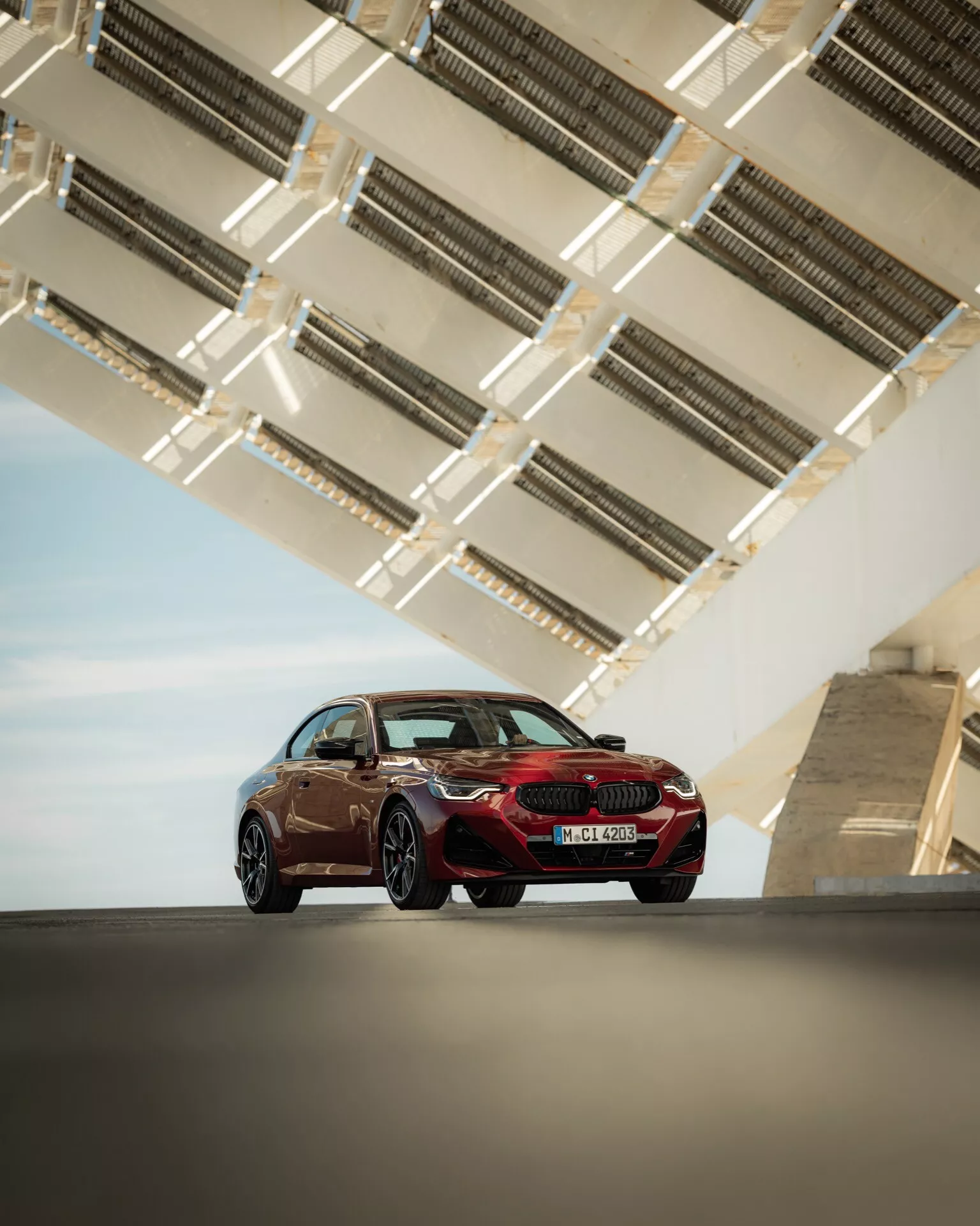 L'image montre une berline sportive de luxe BMW Série 2 2024 (facelift G42-G87) vue en contre-plongée, mettant en valeur sa stature et ses lignes de design. Capturée sous un angle trois-quarts avant, elle montre le côté conducteur du véhicule, exposant la calandre avant, les phares à LED, le contour du capot, le profil latéral, les roues et les portes. La berline est positionnée sur une zone pavée élégante avec une structure architecturale moderne et géométrique au-dessus, probablement une partie d'un pont ou d'une autoroute surélevée, avec des éléments métalliques qui accentuent le design et la couleur de la voiture. La silhouette aérodynamique, les jantes sportives de grand diamètre et les rétroviseurs extérieurs profilés suggèrent une avancée aérodynamique et une technologie moderne du véhicule.
