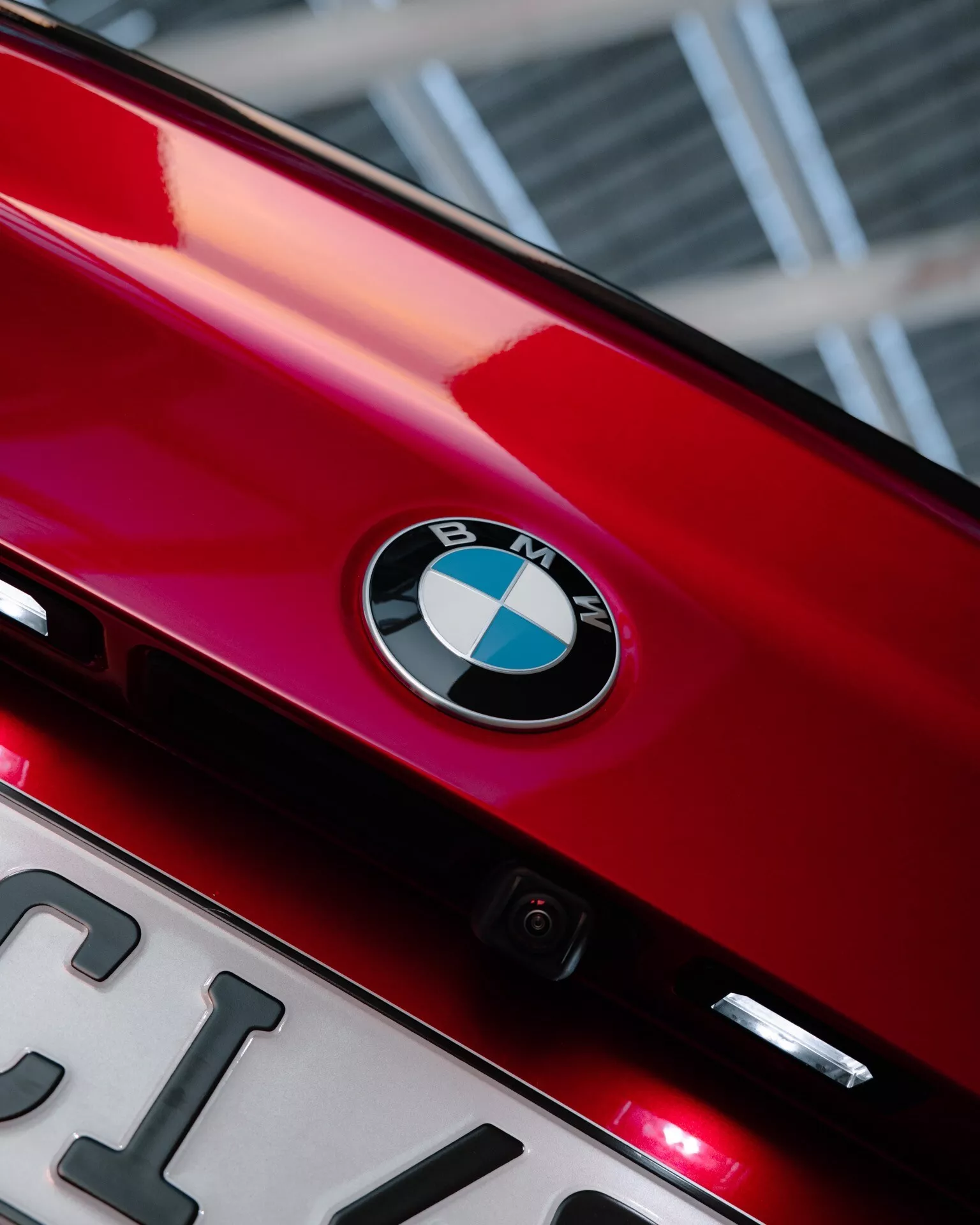 Vue rapprochée de la partie arrière d'une BMW Série 2 2024 rouge brillant (facelift G42-G87), mettant en avant le badge BMW situé au centre sur le couvercle du coffre. Sous l'emblème, une plaque d'immatriculation partiellement visible et une petite caméra arrière mettent en évidence les fonctionnalités technologiques. Deux petites lumières rouges sous le badge pourraient être des feux de freinage supplémentaires ou des feux de brouillard arrière. L'angle de la photo légèrement vers le haut et le cadrage serré mettent en avant l'identité de la marque et les éléments de design élégant, avec un effet bokeh en arrière-plan.