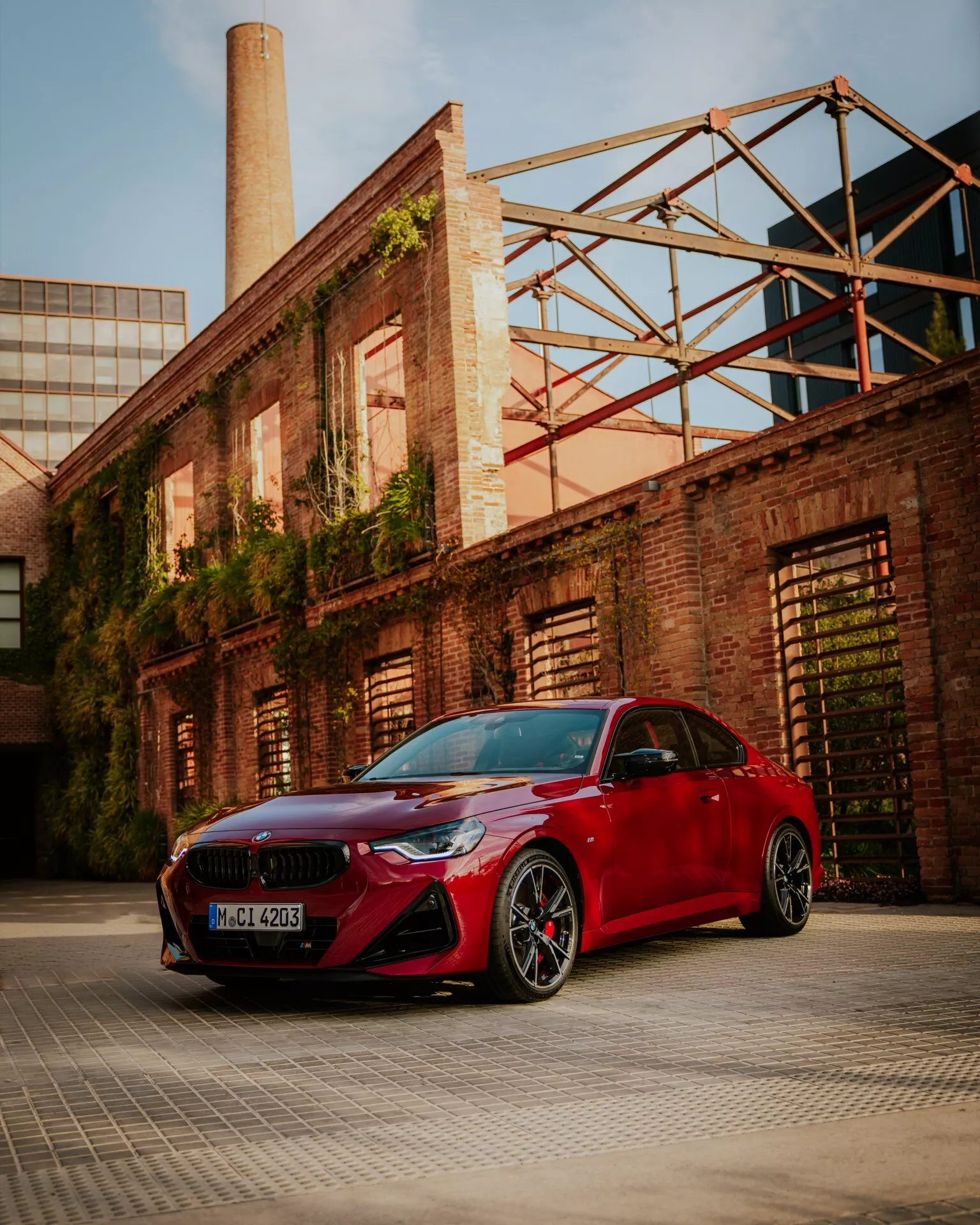 Un angle bas d'une BMW Série 2 2024 rouge (G42-G87-facelift) stationnée dans un quartier industriel ou d'entrepôt, montrant son pare-chocs avant sportif avec de grandes prises d'air, des phares étroits et élégants, une grille en forme de rein et des rétroviseurs latéraux aérodynamiques. L'image montre également des roues en alliage noir à plusieurs rayons avec des pneus à profil bas et des étriers de frein visibles. Les lignes sculptées du capot et la finition de peinture brillante sont accentuées par une lumière ambiante chaude, mettant en valeur les éléments de design luxueux et orientés vers la performance de la voiture.