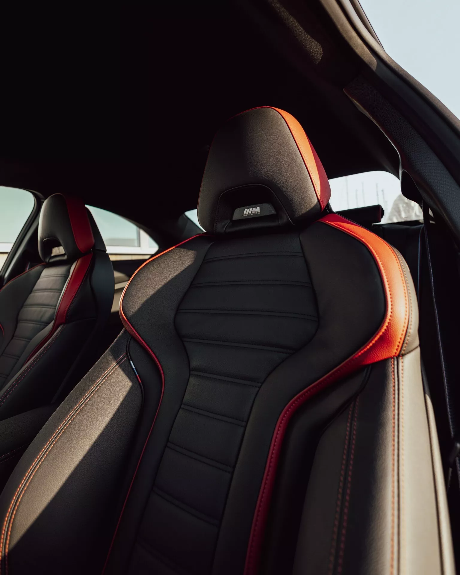 Vue rapprochée du siège passager avant de la BMW 2er-Reihe 2024 (G42-G87 Facelift) depuis le compartiment passager arrière, montrant le luxueux rembourrage en cuir noir avec des coutures rouges et des passepoils rouges. Conception ergonomique avec des renforts latéraux proéminents, un appui-tête brodé avec un logo de sous-marque de luxe ou de performance, le tout à la lumière naturelle.