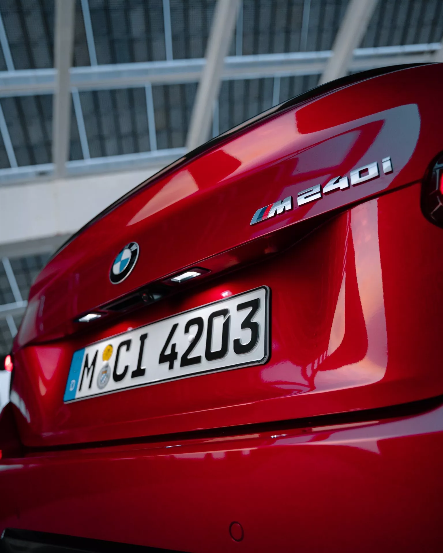 Vue rapprochée de l'arrière d'une BMW M240i rouge, montrant l'emblème BMW, la désignation du modèle, le couvercle du coffre, les feux arrière en forme de L et le spoiler arrière intégré, avec un arrière-plan urbain ou industriel.