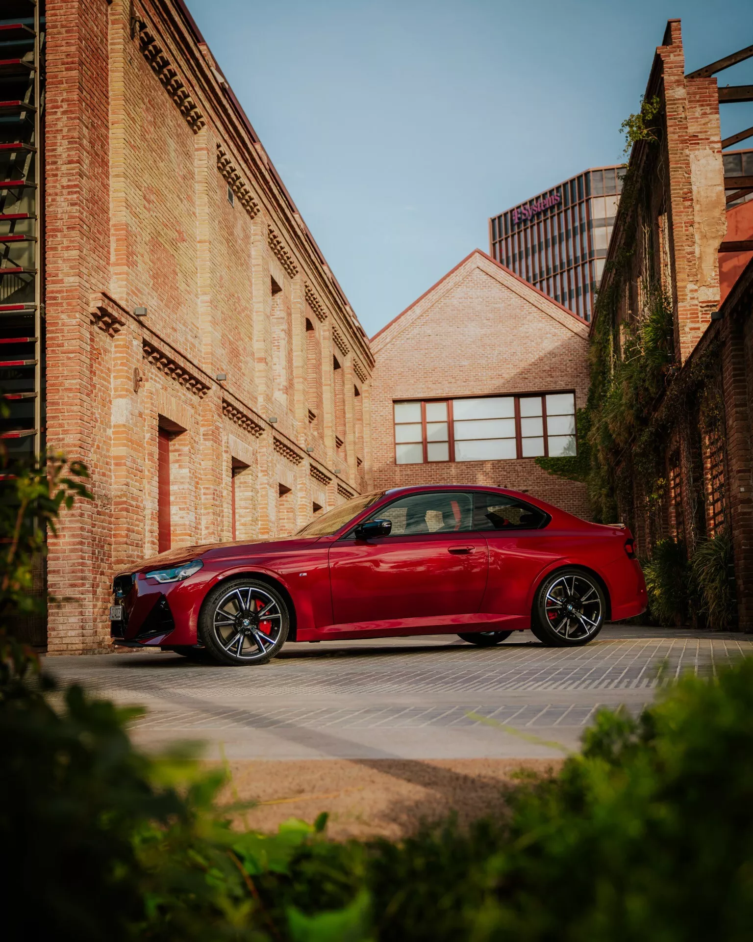 Une BMW Série 2 2024 (facelift G42-G87) rouge garée dans un décor urbain, avec des bâtiments en briques en arrière-plan. La voiture est montrée de profil avec des lignes de design sur les portes et les ailes, et des jantes en alliage sportives clairement visibles. L'image est prise sous un angle bas, améliorant la posture imposante du véhicule contre le soleil du matin ou de l'après-midi qui projette des ombres douces. Cette photo capture des thèmes de design automobile, de finitions sportives et de style de vie urbain.
