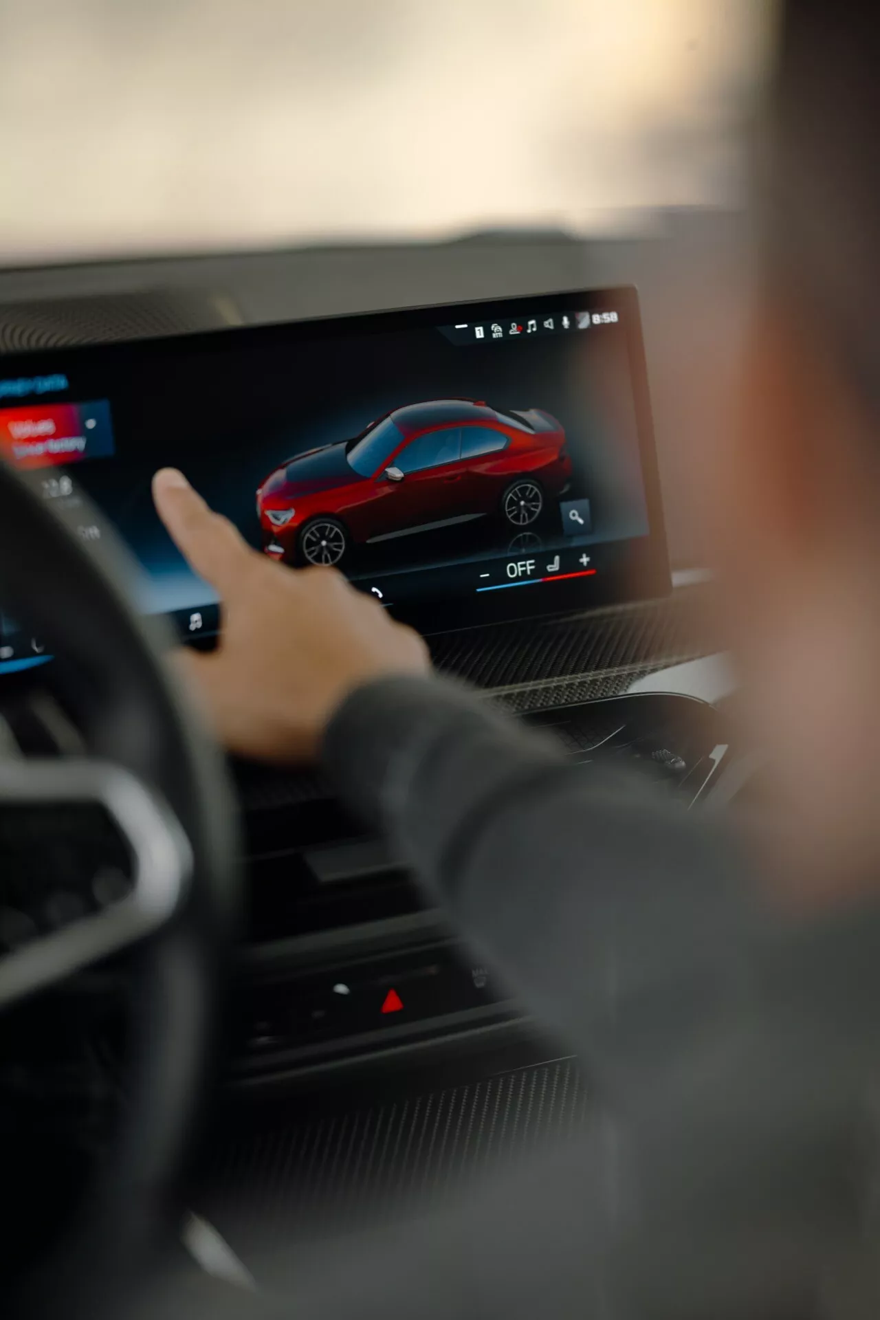 Vue rapprochée d'un système d'infodivertissement à écran tactile moderne en voiture vu depuis le siège du conducteur, se concentrant sur une interface de personnalisation affichant une voiture de sport rouge. Le volant est légèrement flou, mettant en évidence l'installation technologique avancée de la BMW Série 2 2024 G42-G87 restylée avec des fonctionnalités avancées pour la navigation, les réglages, la connectivité et le divertissement multimédia.