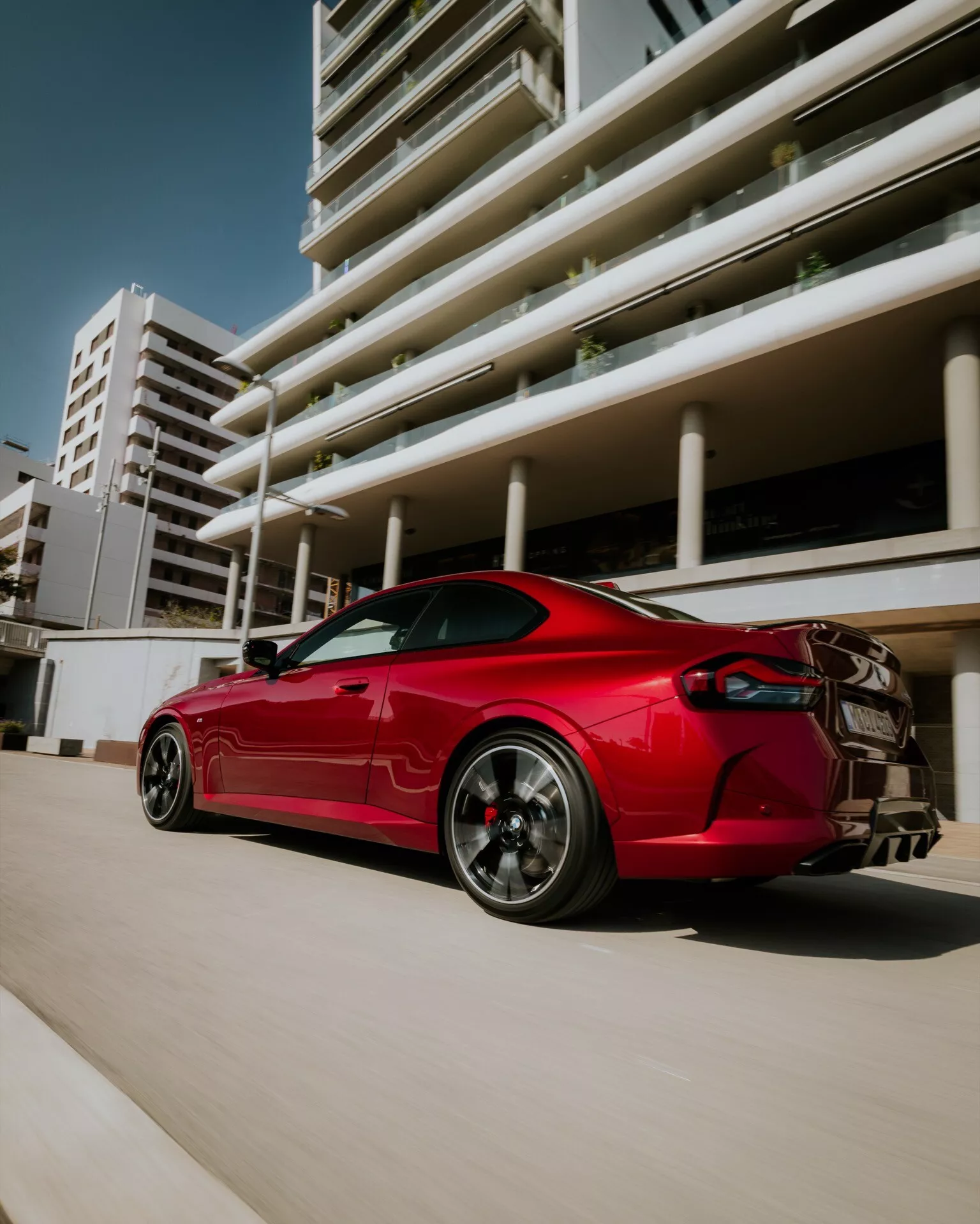Photographie de la BMW Série 2 2024 (Facelift G42-G87) en rouge vibrant, présentant un élégant coupé sportif d'une perspective arrière trois quarts basse. Caractéristiques incluent des lignes aérodynamiques, des jantes en alliage multi-branches graphite avec des étriers de frein de performance visibles, un aileron arrière proéminent, un système d'échappement double et des feux arrière LED nets. L'image capture la voiture dans un environnement urbain avec des bâtiments modernes en arrière-plan, soulignant son allure agressive et son design sportif.