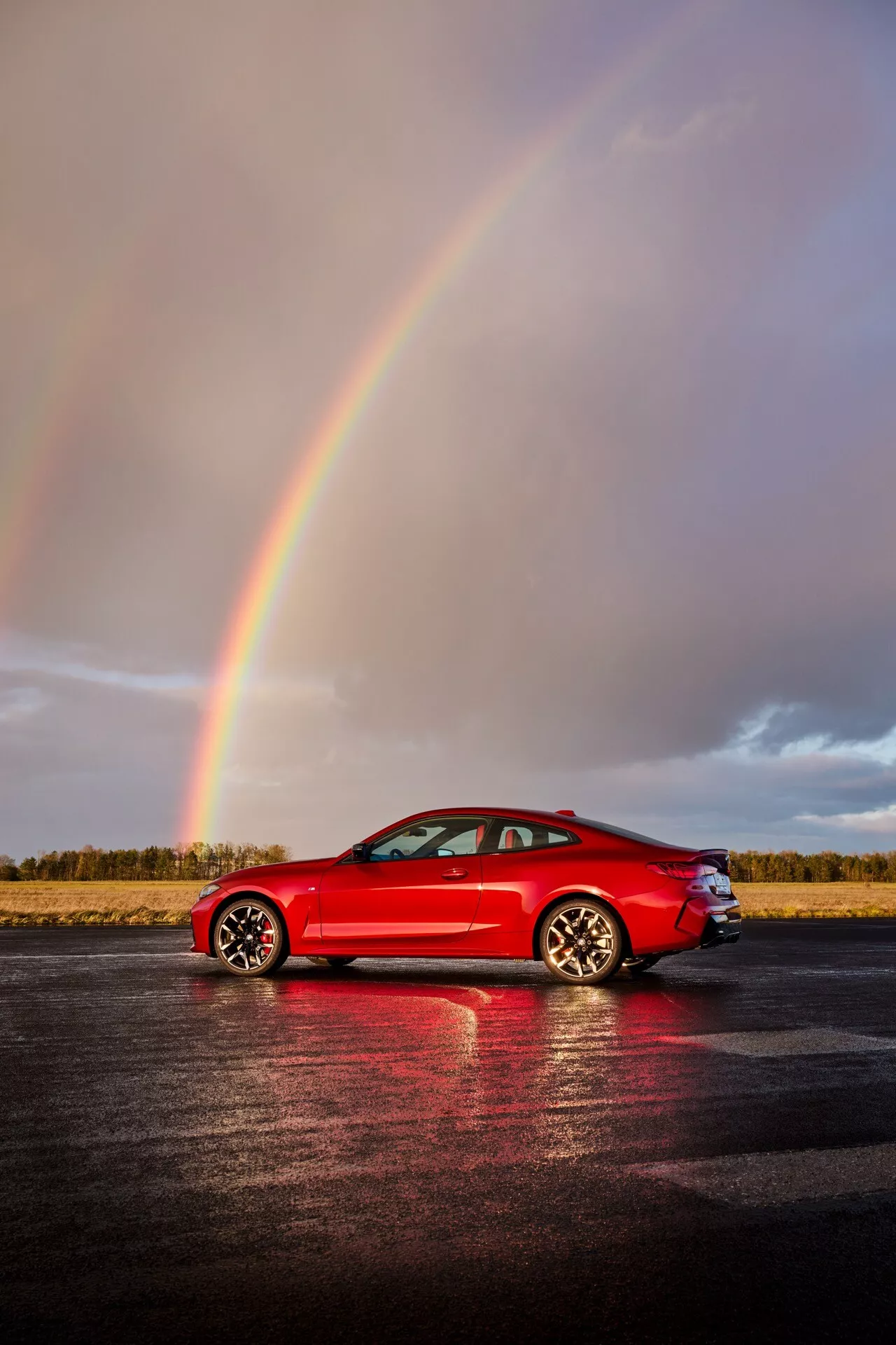 Coupé sportif rouge BMW Série 4 2024 (G22-G23-G26-G82-G83 Facelift) garé sur un asphalte mouillé réfléchissant sous un arc-en-ciel