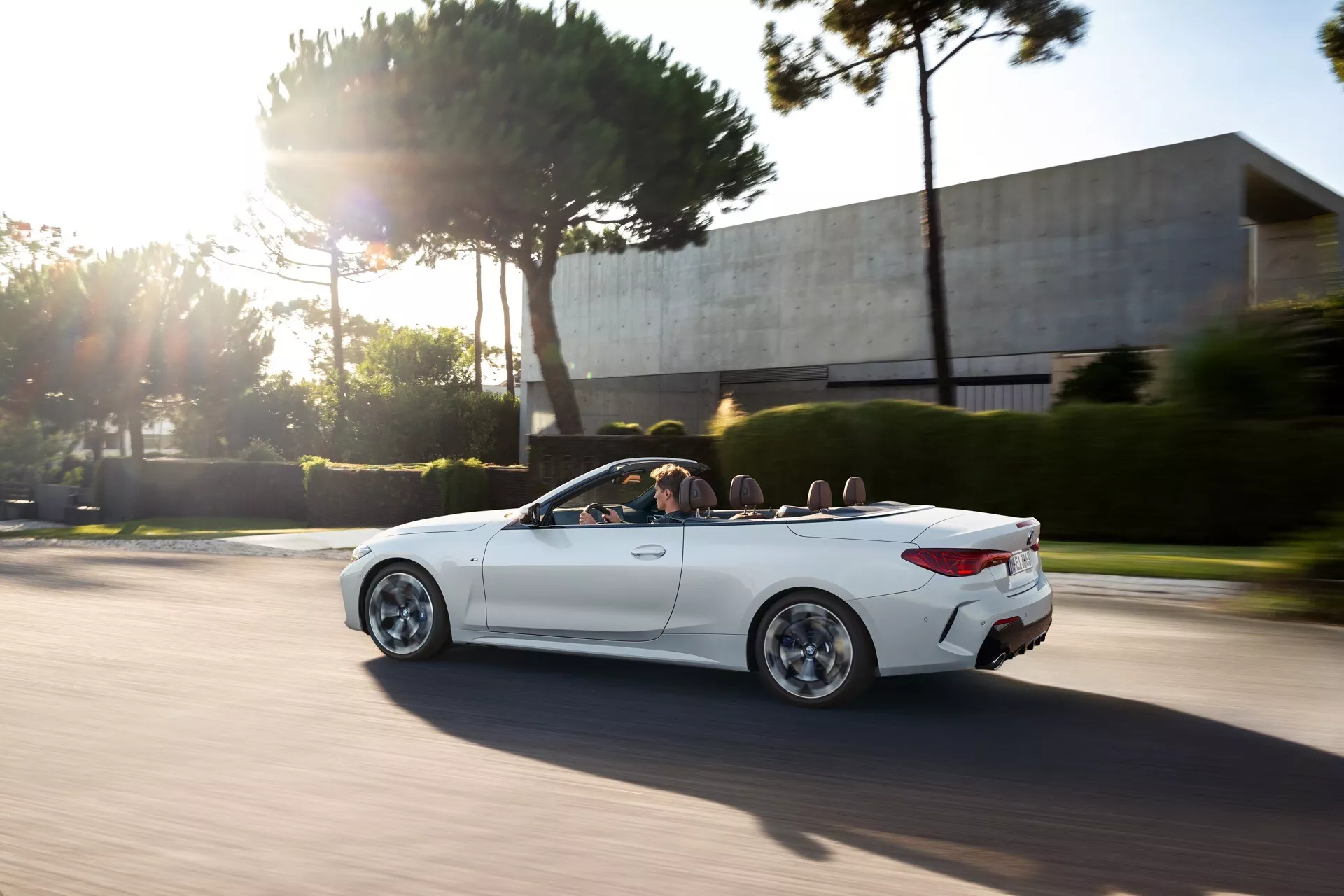 BMW Série 4 Cabriolet (G22-G23-G26-G82-G83 Facelift) 2024 en mouvement, montrant son design élégant blanc avec des passages de roue proéminents, des roues en alliage stylées et une vue claire sur son intérieur luxueux à travers le mécanisme de toit convertible, pendant l'heure dorée avec une posture sportive dynamique soulignée par le flou de vitesse et l'éclat du soleil.