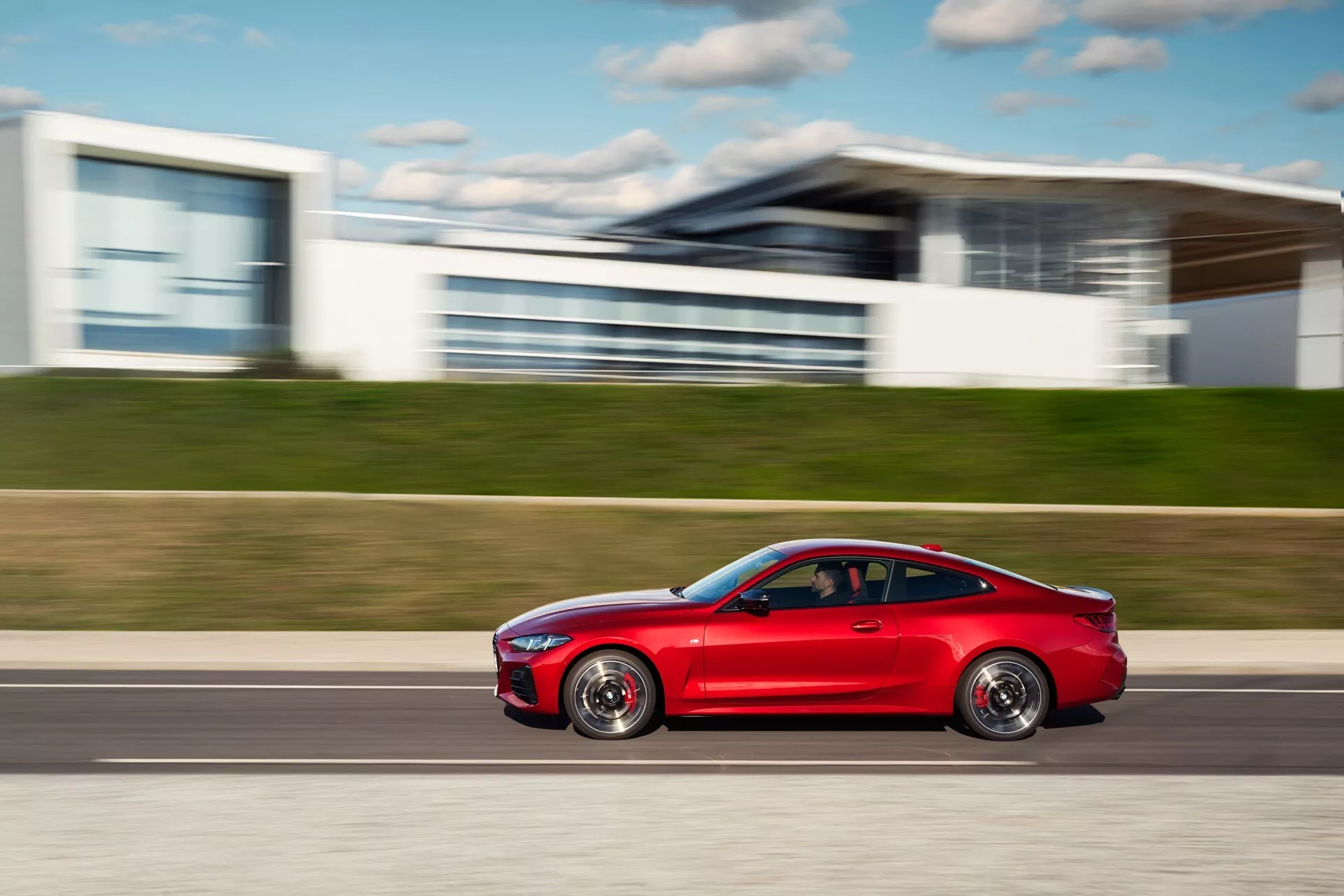 Coupé rouge moderne BMW Série 4 (G22-G23-G26-G82-G83 Facelift) en mouvement, affichant un design aérodynamique, des jantes alliage sportives, des vitres teintées et des pneus hautes performances, capturé dans une prise de vue latérale dynamique sur un fond urbain flou.
