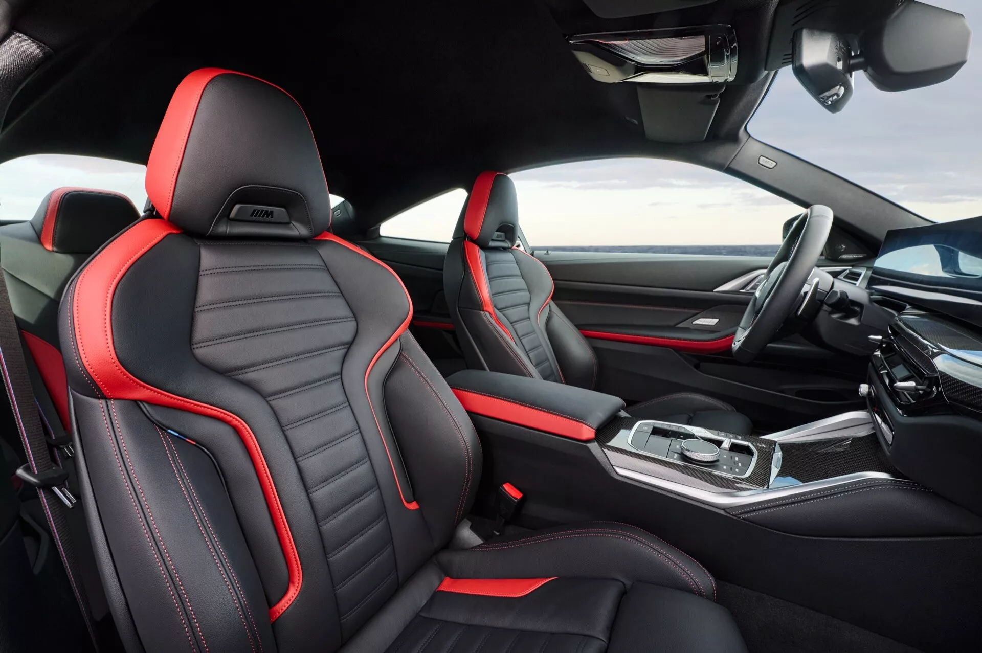 Vue intérieure de la BMW Série 4 2024 (G22-G23-G26-G82-G83 Facelift) mettant en avant les sièges avant sportifs en cuir noir avec coutures rouges, console centrale élégante avec accents métalliques, affichage avancé du tableau de bord d'infodivertissement et design ergonomique moderne