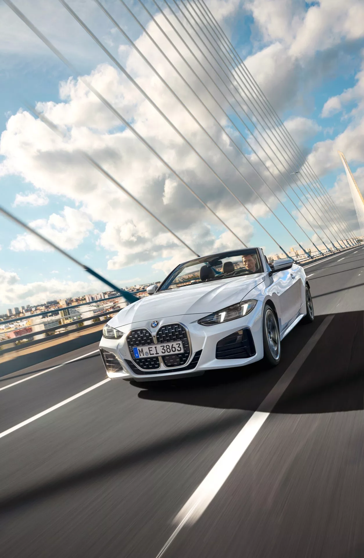 Convertible BMW Série 4 2024 (lifting G22-G23-G26-G82-G83) en blanc, capturée dynamiquement d'un angle avant bas en traversant un pont suspendu sous un ciel partiellement nuageux, démontrant son design élégant, éclairage LED et fonctionnalités aérodynamiques