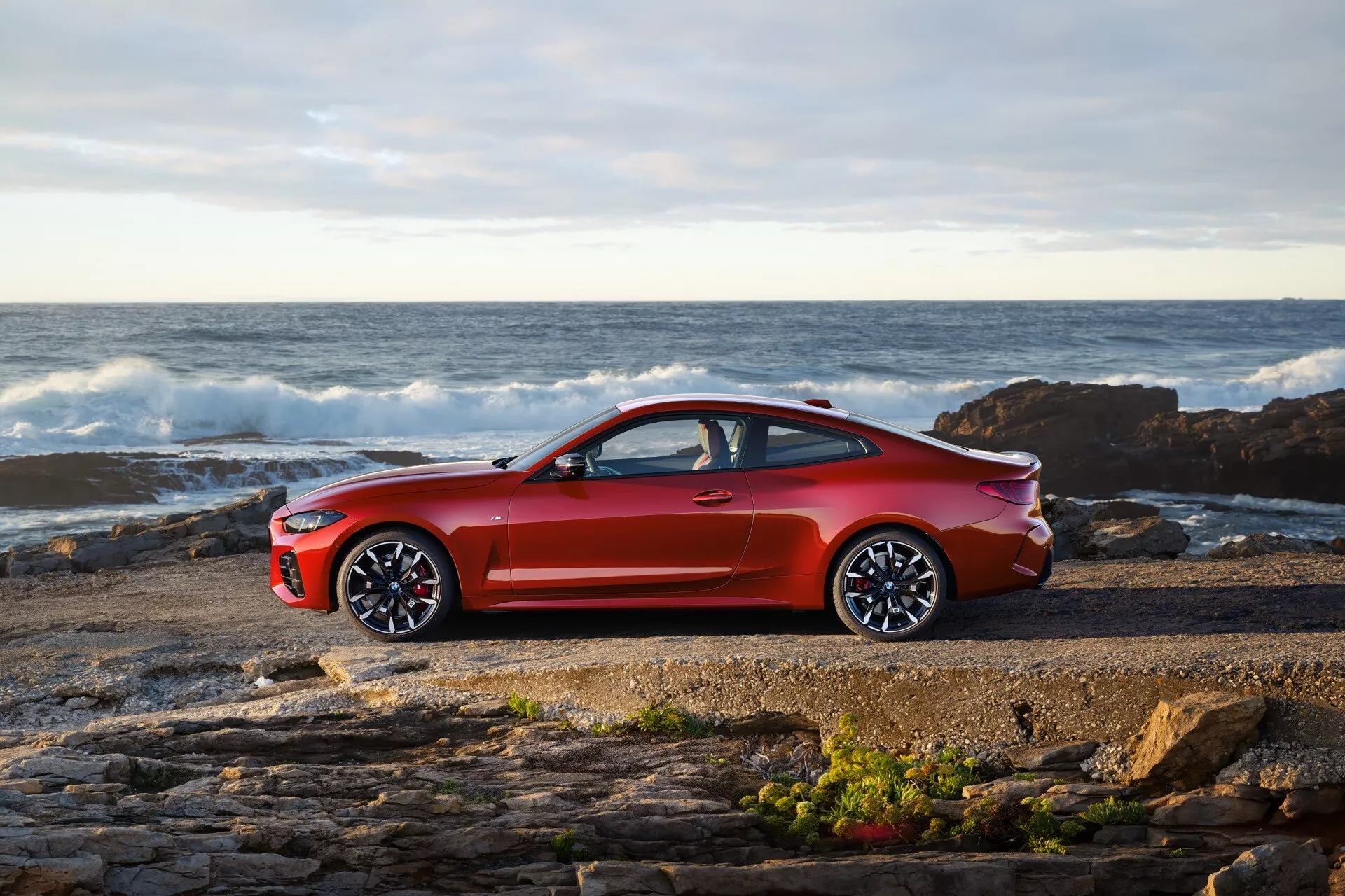 Coupé BMW Série 4 (G22-G23-G26-G82-G83 Facelift) 2024 en rouge métallique brillant garée près d'un rivage rocheux avec les vagues et l'océan en arrière-plan sous un ciel partiellement nuageux, présentant son profil latéral sportif avec de grandes jantes alliage à multiples branches et un design aérodynamique élégant