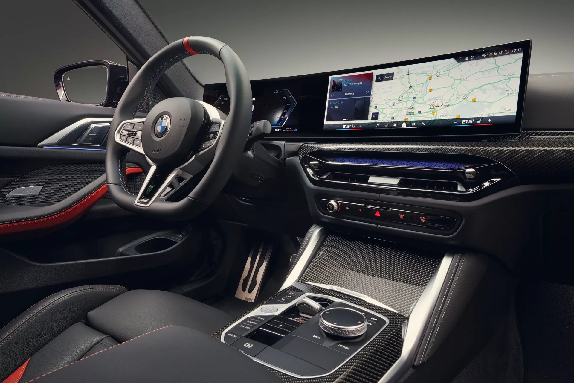 Vue intérieure de la BMW Série 4 2024 (G22-G23-G26-G82-G83 Facelift) montrant le badge sur le volant sportif multifonction, le tableau de bord numérique avec affichage allongé, la console centrale élégante en finition brillante et les sièges avec un schéma de couleurs bicolore avec des accents rouges.
