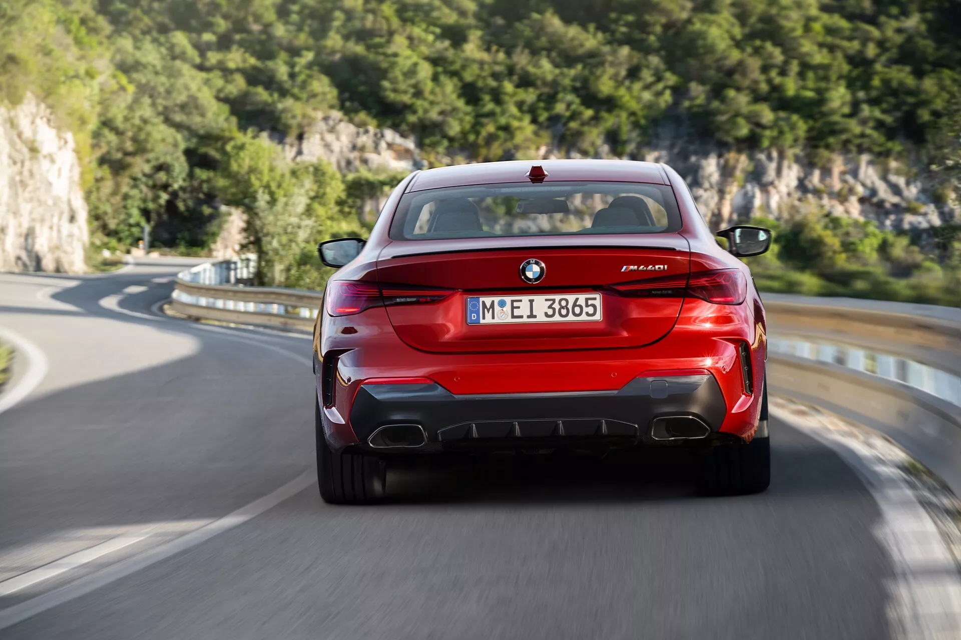 Vue arrière d'une BMW M8 Competition rouge en mouvement, affichant l'emblème BMW, les badges M8 et de la série M, des feux arrière distinctifs, quatre tuyaux d'échappement et un diffuseur arrière aérodynamique sur une route de campagne avec un décor naturel en arrière-plan.