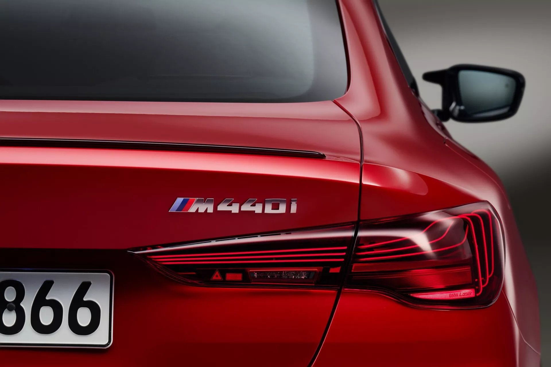 Vue rapprochée et angulaire du quart arrière d'une BMW Série 4 2024 (G22-G23-G26-G82-G83-Facelift) M440i rouge, mettant en vedette le badge du modèle, des feux arrière LED élégants, un design extérieur sculpté et une finition rouge métallique brillante sous un éclairage artificiel.