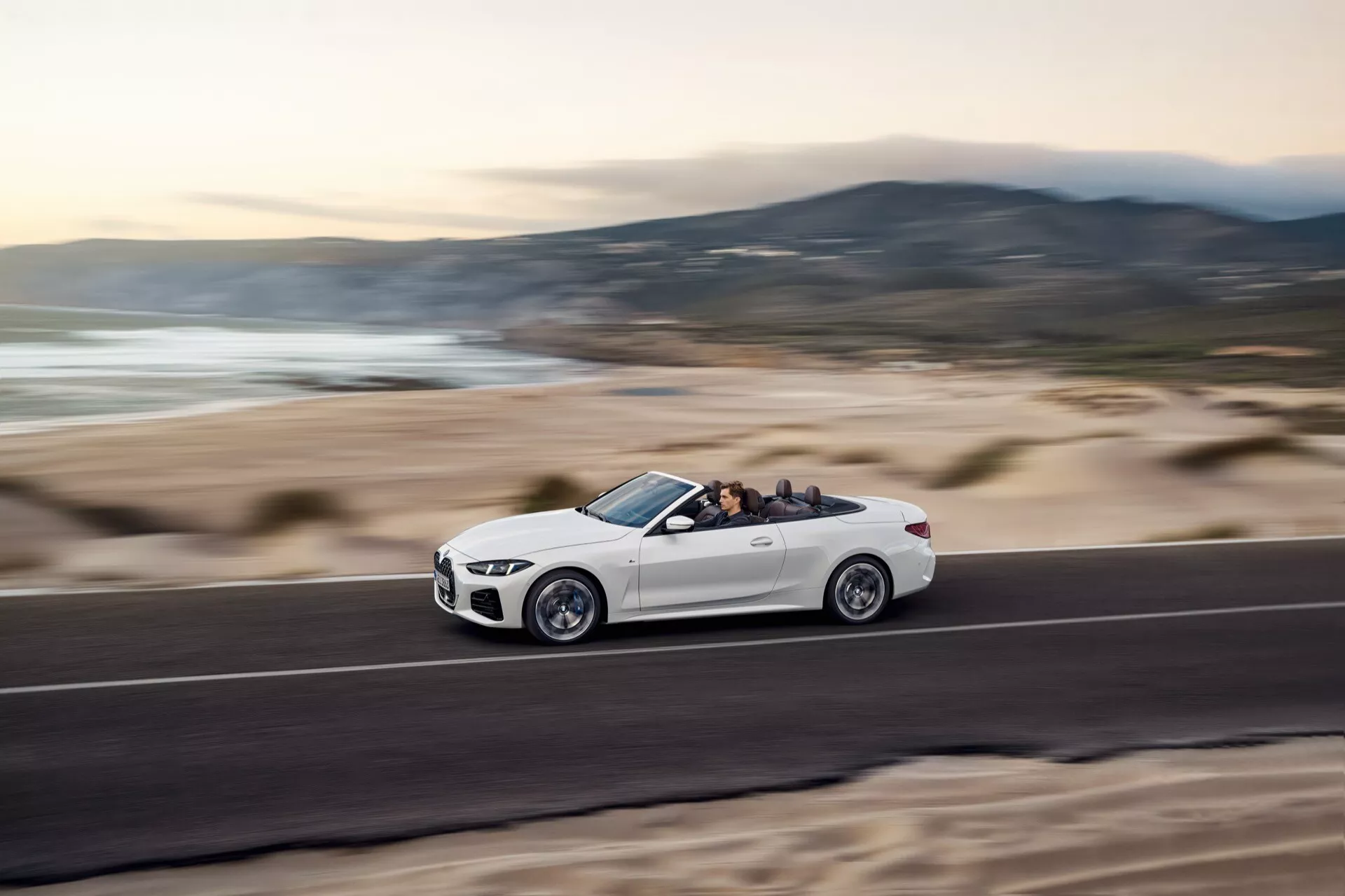 BMW Série 4 Cabriolet 2024 (G22-G23-G26-G82-G83 Facelift) en mouvement sur une route côtière, montrant le profil latéral avec une posture dynamique grâce à la prise de vue en angle bas, détails des roues et finitions visibles, toit ouvert révélant l'intérieur et les occupants, sur fond de paysage de plage flou