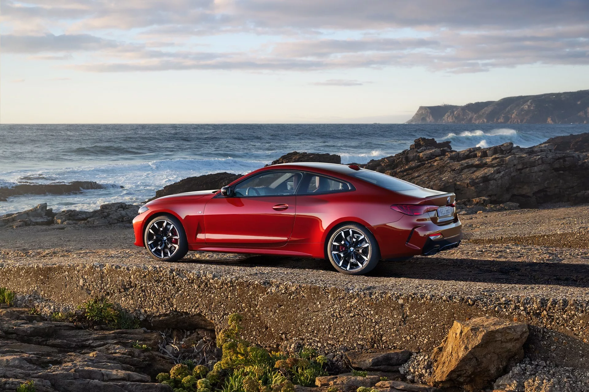 Coupé sportif moderne BMW Série 4 2024 (G22-G23-G26-G82-G83-Facelift) en rouge métallisé garé sur une côte rocheuse avec l'océan en arrière-plan, montrant le profil latéral et le design arrière incluant les feux arrière, le pare-chocs, les roues en alliage et les éventuels tuyaux d'échappement depuis une perspective arrière de trois quarts.