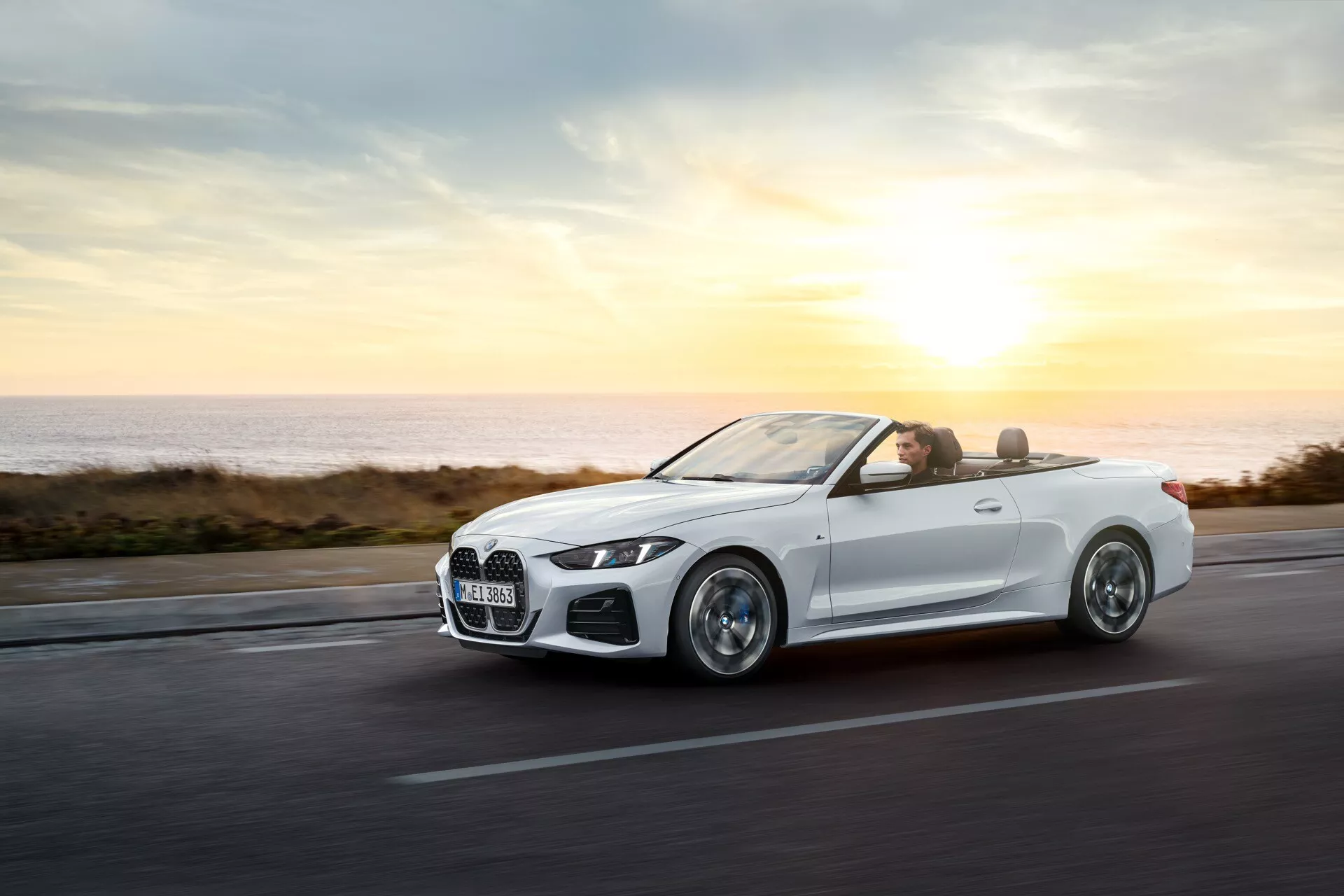 BMW Série 4 Cabriolet (G22-G23-G26-G82-G83 Facelift) 2024 en mouvement sur une route côtière à l'aube ou au crépuscule, exhibant sa carrosserie aérodynamique, ses roues en alliage, ses phares LED, son intérieur luxueux et sa technologie avancée