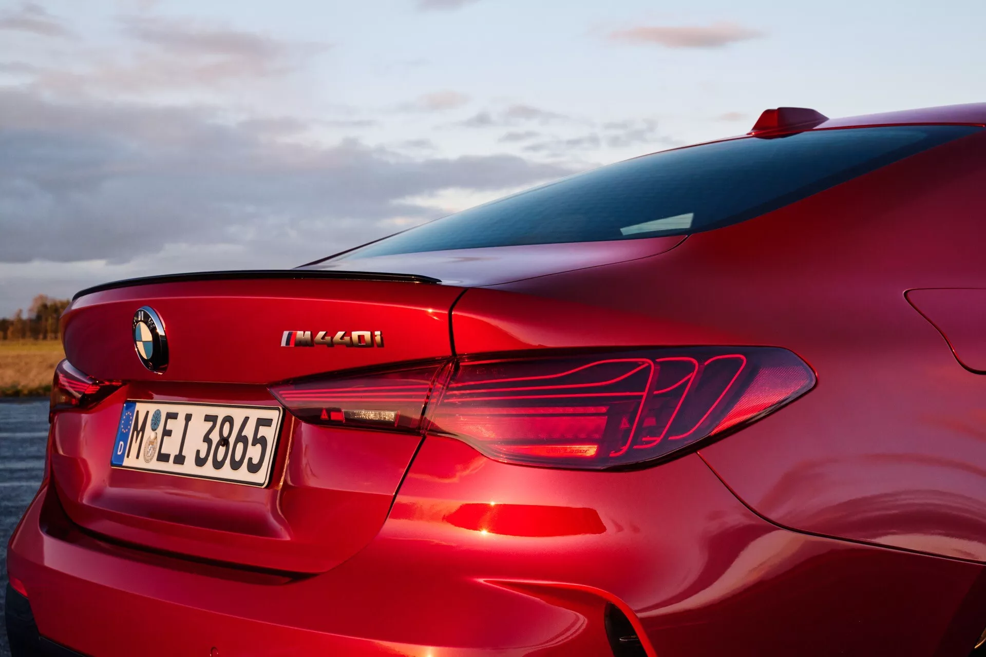 Section arrière de la BMW Série 4 2024 (G22-G23-G26-G82-G83 Facelift) vue sous un angle haut de trois quarts, présentant le panneau arrière droit, le design des feux arrière LED, le diffuseur arrière intégré, une partie de la vitre arrière et du pilier C, avec un focus sur les éléments de design de la berline sportive de luxe, le badging de performance, et la finition rouge métallique dans un cadre extérieur au lever ou coucher du soleil.