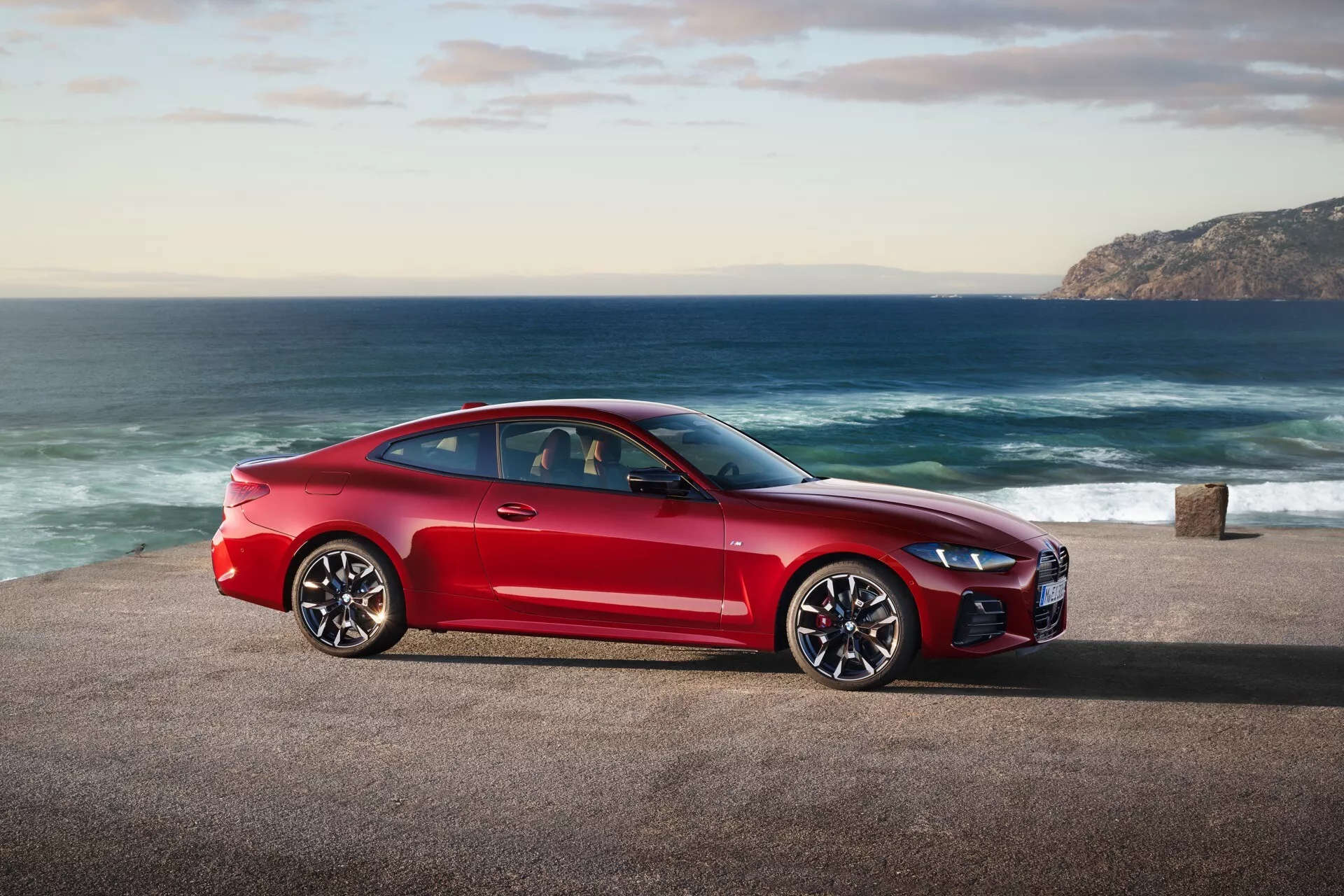 Coupé sportif rouge BMW série 4 (facelift G22-G23-G26-G82-G83) 2024 garé sur une route côtière avec un océan et côte vallonnée en arrière-plan, montrant le profil latéral du véhicule, un design élégant et des caractéristiques de performance à la lumière naturelle du jour