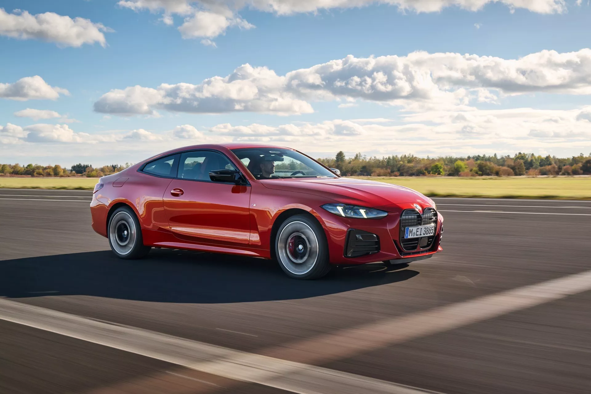 Modèle restylé de la BMW Série 4 (G22-G23-G26-G82-G83) 2024 en rouge, montrant un mouvement dynamique et un design sportif avec une forme de coupé basse, de grandes roues en alliage et une silhouette aérodynamique sur une piste d'atterrissage sous un ciel clair