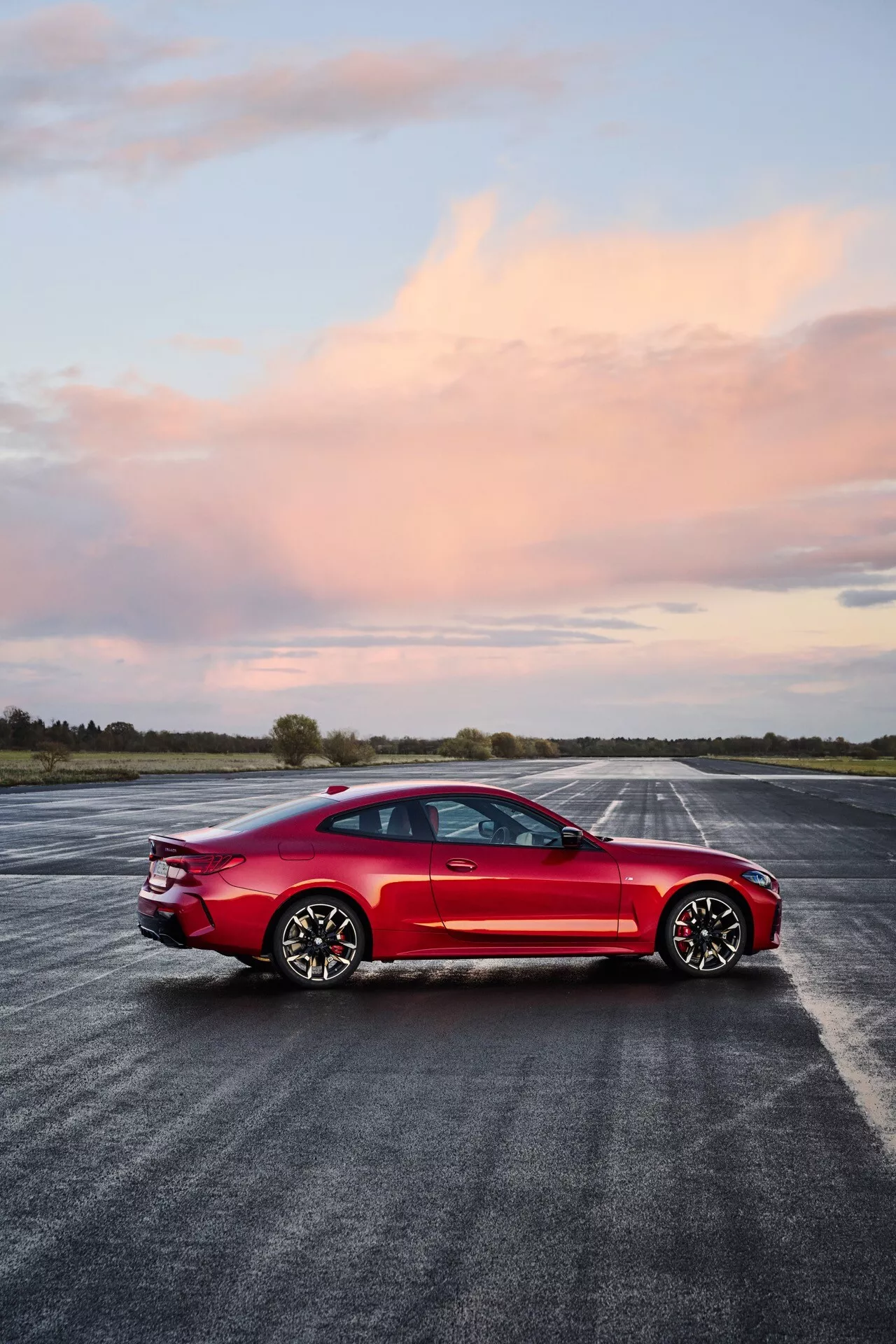 Berline sportive rouge quatre portes BMW Série 4 (G22-G23-G26-G82-G83 Facelift) sur tarmac mouillé, montrant le profil latéral, design aérodynamique, phares LED, toit panoramique, jantes alliage multi-rayons et freins haute performance sous un ciel au coucher du soleil