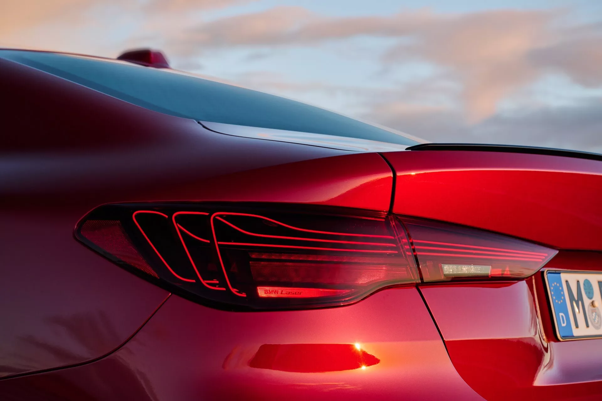Gros plan sur le feu arrière gauche de la BMW Série 4 2024 (G22-G23-G26-G82-G83 restylée) avec bandes lumineuses LED horizontales et design de nageoires aérodynamiques, sur fond de ciel au coucher du soleil