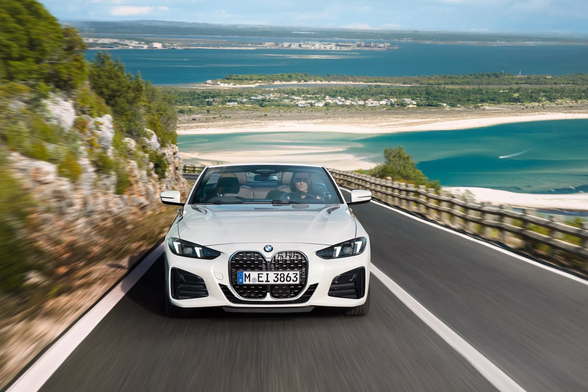 Berline de luxe BMW Série 4 (G22-G23-G26-G82-G83 facelift) de 2024 roulant le long d'une route côtière pittoresque, affichant sa grande calandre en forme de rein, ses phares LED inclinés et son design dynamique de pare-chocs avant