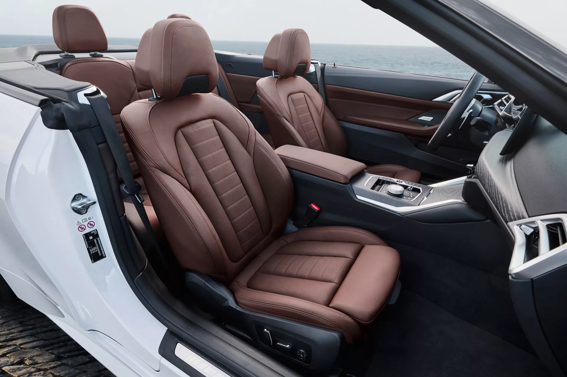 Intérieur de la BMW Série 4 2024 (G22-G23-G26-G82-G83 lifting) mettant en avant la cabine avant depuis une perspective arrière-côté, soulignant les sièges en cuir marron luxueux, la nature convertible et les commandes avancées sur la porte et la console centrale.
