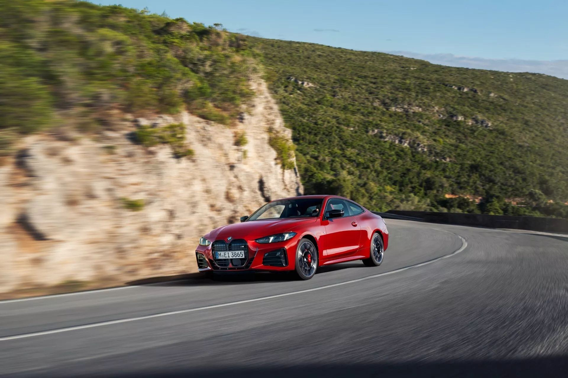 Vue dynamique de l'angle avant latéral d'une BMW Série 4 2024 (G22-G23-G26-G82-G83 facelift) en rouge, présentant son style agressif, ses lignes aérodynamiques, son fascia avant sportif, son capot, son panneau latéral, sa roue avant et son pneu sur une route sinueuse