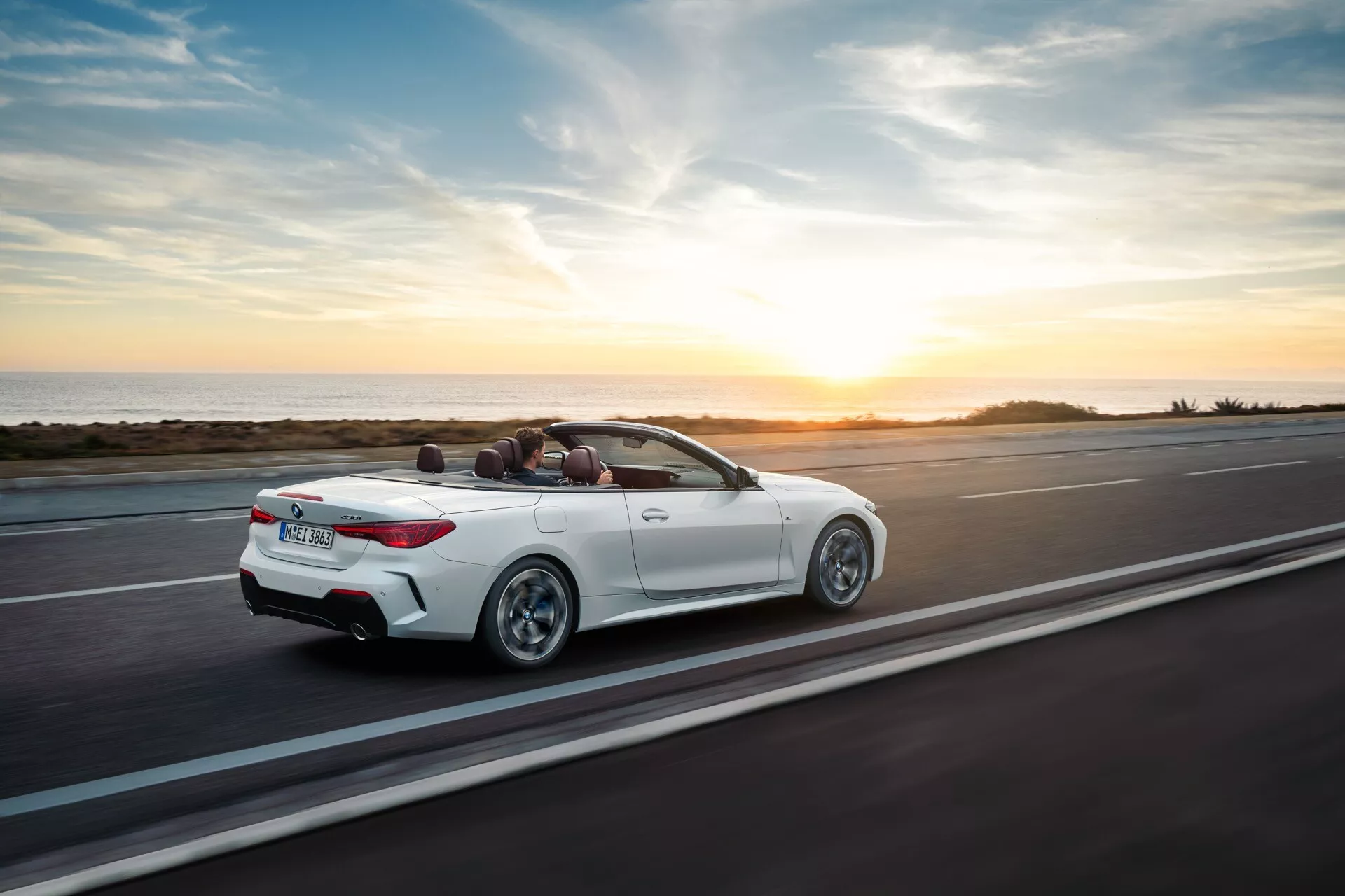 BMW Série 4 Cabriolet (G22-G23-G26-G82-G83 restylé) en mouvement sur une route côtière au coucher du soleil, vue arrière aérienne trois quartiers montrant son extérieur blanc, toit convertible baissé et design sportif
