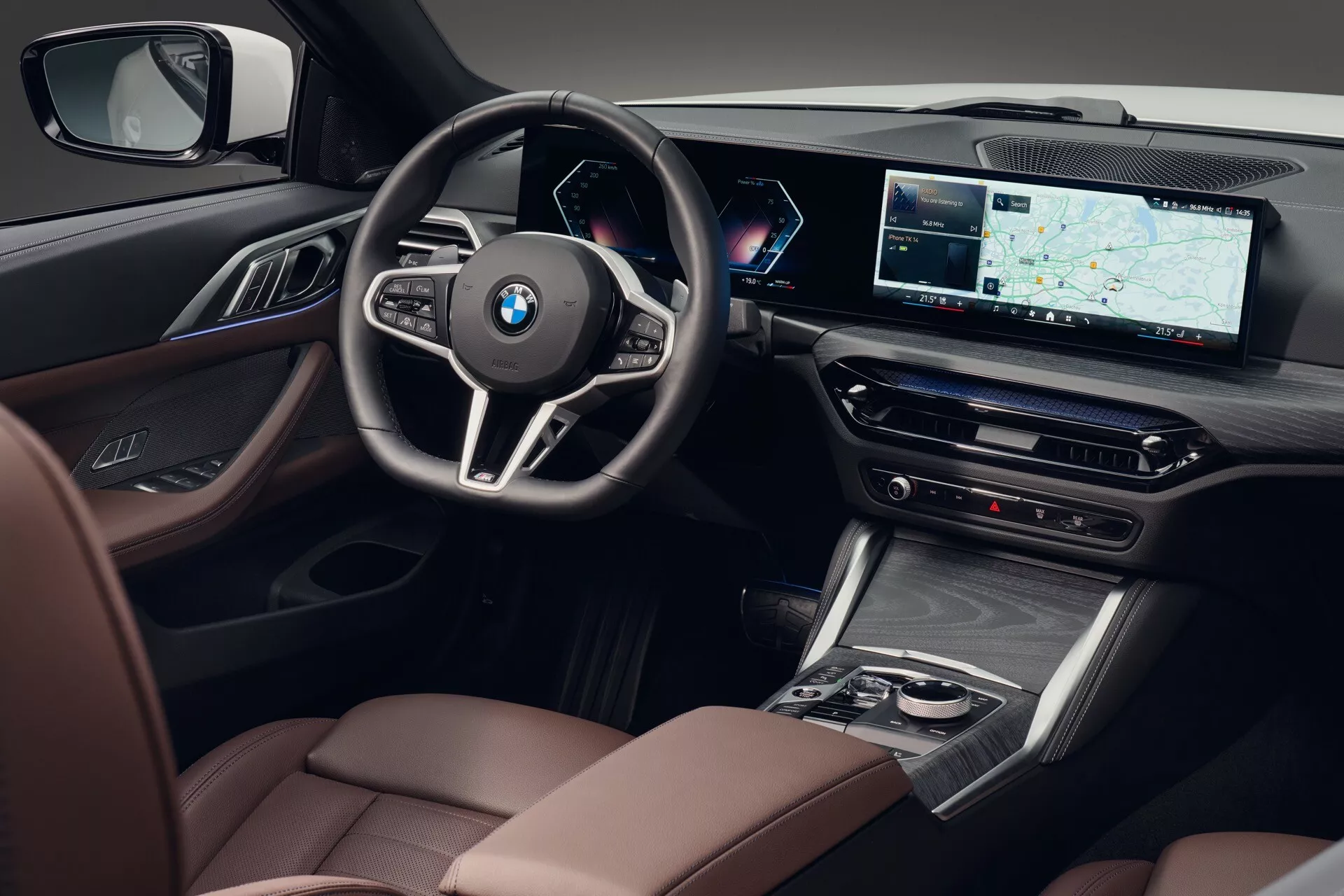 Vue intérieure depuis un angle légèrement élevé se concentrant sur le côté conducteur de la BMW Série 4 2024 (G22-G23-G26-G82-G83 facelift), mettant en avant une sellerie en cuir de haute qualité avec des coutures contrastées, des finitions en bois ou effet bois, des commandes du système audio premium sur le panneau de porte, un volant sportif multifonctions à trois branches avec l'insigne BMW, un cluster d'instruments entièrement numérique et personnalisable derrière le volant, un grand écran d'infotainement large avec navigation GPS, des contrôles de climatisation, un élégant levier de vitesse, une console centrale avec un pavé tactile ou un contrôleur entouré de détails chromés, et le siège du passager luxueux visible, soulignant les caractéristiques de technologie et de luxe.