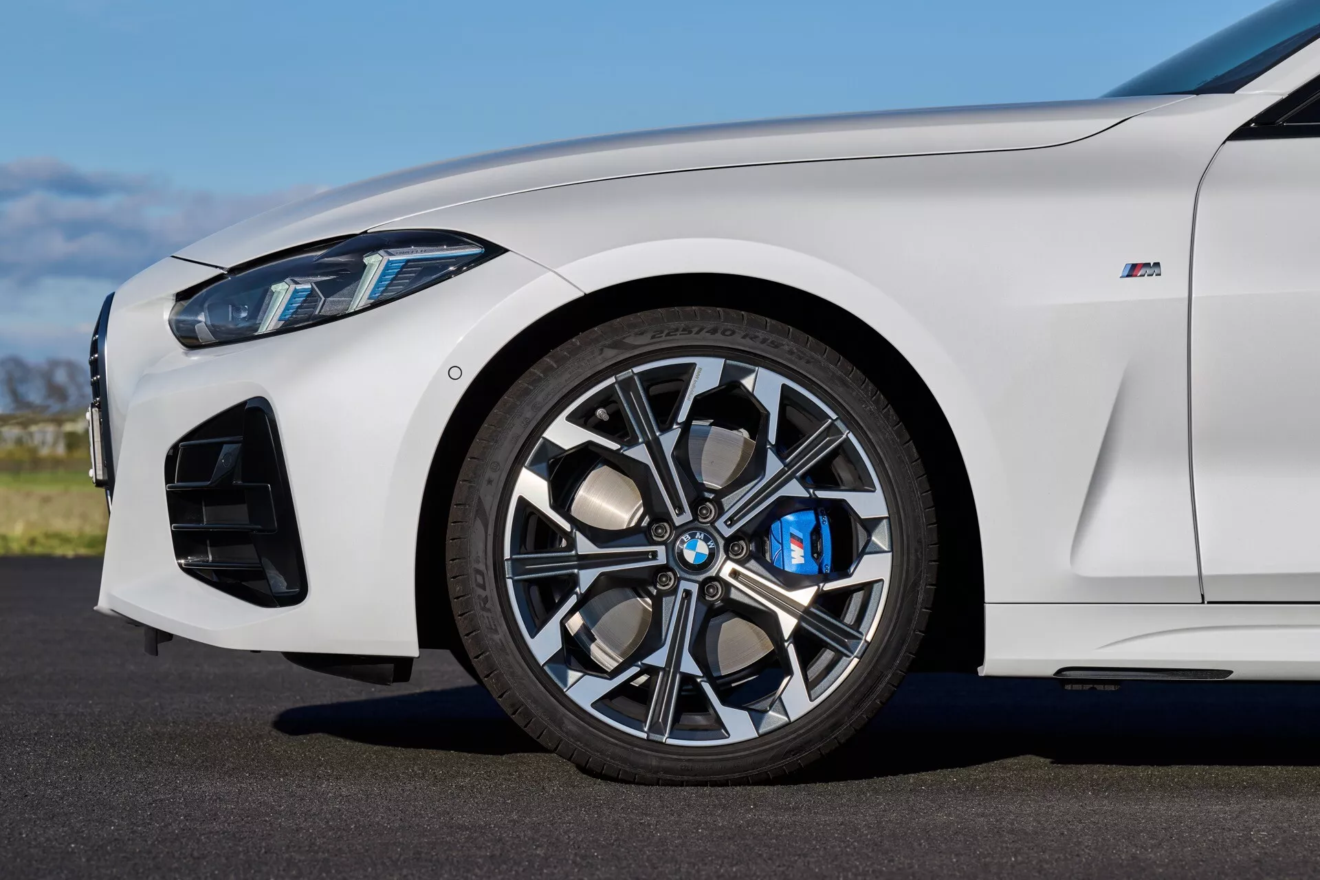 Vue latérale rapprochée de la BMW Série 4 2024 (G22-G23-G26-G82-G83 Facelift) en blanc, se concentrant sur la roue avant avec jante en alliage à multiples rayons, étrier de frein bleu et prises d'air orientées performances sous la lumière naturelle du jour