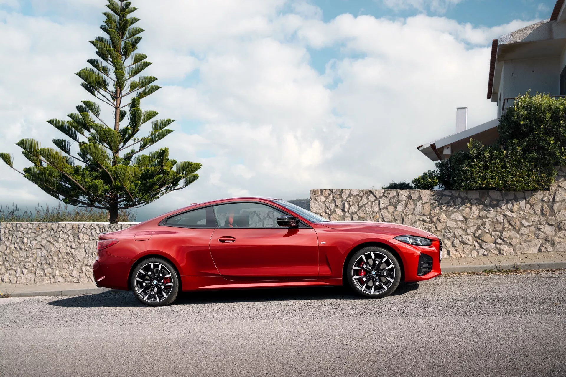 Coupé sportif rouge BMW Série 4 2024 (G22-G23-G26-G82-G83-Facelift) garé sur un trottoir, montrant son profil latéral complet avec une perspective dynamique sous un angle bas soulignant son design sportif, long capot, ligne de toit élégante, grandes roues en alliage, rétroviseurs aérodynamiques, poignées de porte affleurantes, système de freinage performant avec étriers rouges, phares LED et pare-chocs avant agressif, sous un ciel partiellement nuageux à côté de la verdure et d'un bâtiment résidentiel