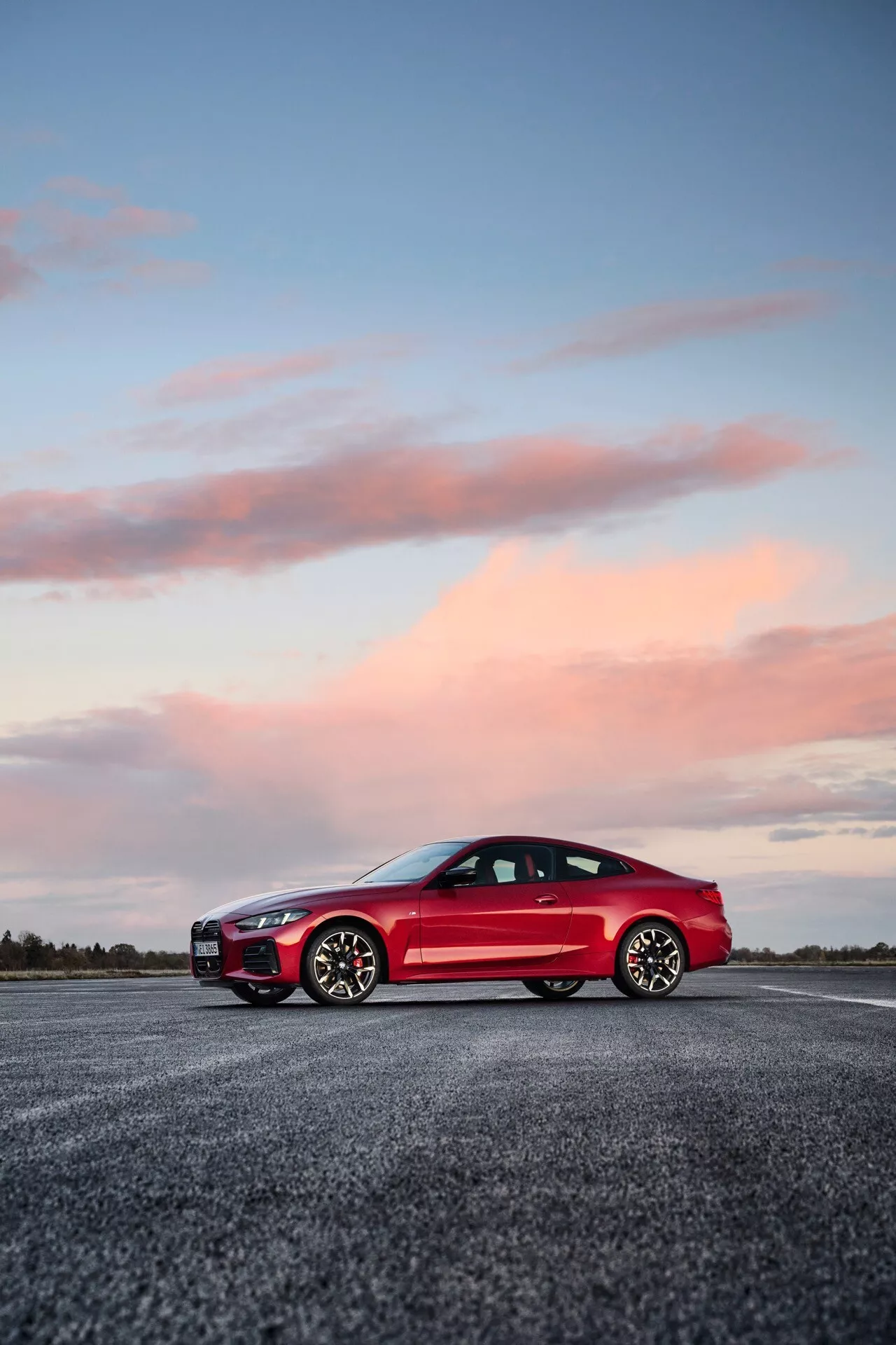 Voiture de sport rouge à deux portes BMW Série 4 2024 (G22-G23-G26-G82-G83 Facelift) mise en valeur sous un angle bas accentuant sa posture, ses lignes de design et ses caractéristiques technologiques telles que les phares LED et les rétroviseurs aérodynamiques, sur un fond de crépuscule