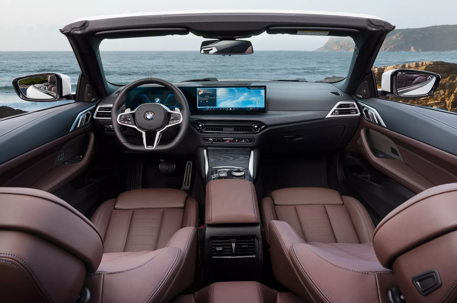 Intérieur du cabriolet BMW Série 4 2024 présentant des sièges avant en cuir marron, un volant à trois branches avec commandes intégrées, un affichage numérique grand écran sur le tableau de bord et une console centrale élégante au sein d'une cabine luxueuse avec le toit convertible rétracté, affichant un décor côtier pittoresque