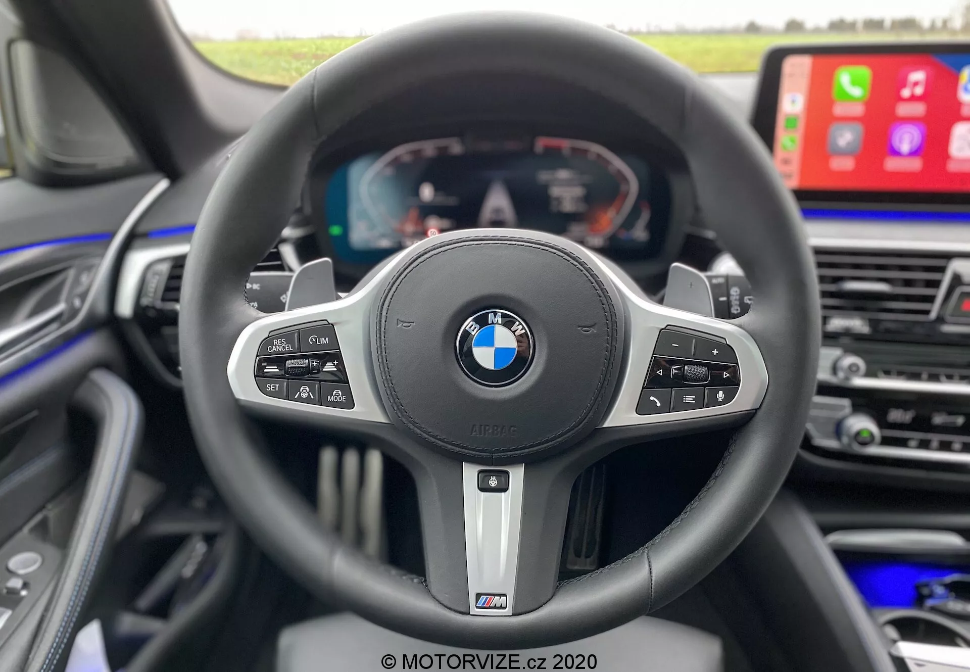 Vue intérieure depuis la perspective du conducteur à l'intérieur d'une BMW 520i Touring Steptronic 2020, mettant en évidence un volant sportif à trois branches avec les badges BMW et M. Le volant comporte plusieurs boutons de commande pour le régulateur de vitesse, les médias, la commande vocale et les fonctions téléphoniques, et des palettes de changement de vitesse indiquant une transmission automatique avec des options manuelles. Derrière le volant, un affichage entièrement numérique montre le compteur de vitesse, le tachymètre et les informations sur le véhicule. À droite, une partie de la console centrale est visible avec un grand écran d'infodivertissement affichant Apple CarPlay, mettant l'accent sur le luxe, la technologie avancée et le design axé sur le conducteur.