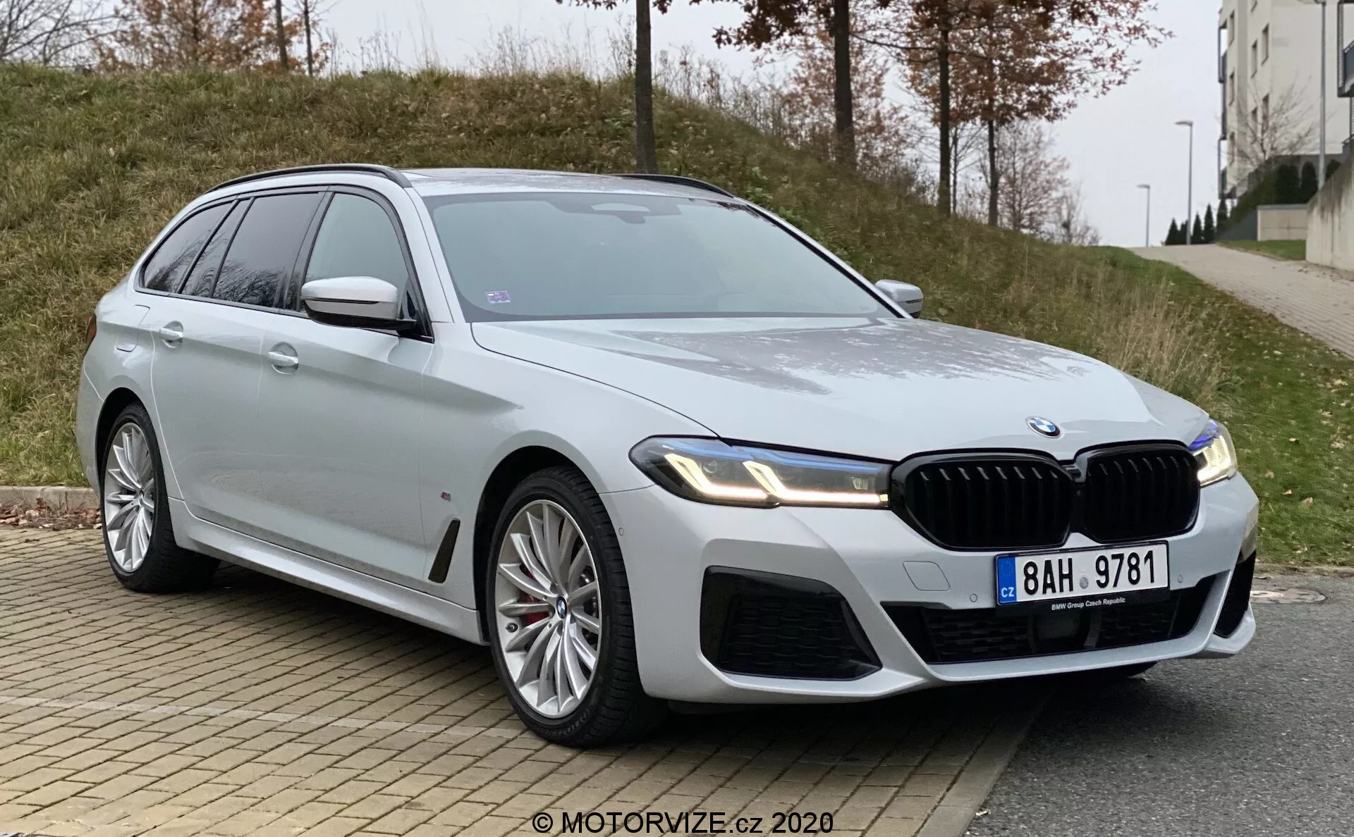 Une BMW Série 5 moderne et luxueuse (BMW 520i Touring Steptronic) break photographiée sous un angle de trois quarts avant bas. L'image met en valeur la calandre avant de la voiture, les phares angulaires et élégants probablement dotés de la technologie LED, et les lignes de conception musclées sur le capot et les ailes. Les parties visibles incluent la calandre avant, le pare-chocs, les phares, la porte avant côté conducteur, l'aile avant, une partie de la porte arrière, les fenêtres latérales, les roues et le miroir latéral côté conducteur. Les grandes jantes en alliage suggèrent une variante sportive du modèle, et la peinture argentée est réfléchissante et propre. La voiture présente une calandre avant proéminente avec des lattes verticales, un badge sur l'aile avant indiquant un niveau de finition supérieur, et des rétroviseurs latéraux avec clignotants intégrés. Un toit ouvrant est également visible. Le véhicule est garé sur un bord de route urbain avec un trottoir et un pavé au premier plan et une zone de pelouse et un bâtiment à l'arrière-plan, sous des conditions d'éclairage nuageuses.