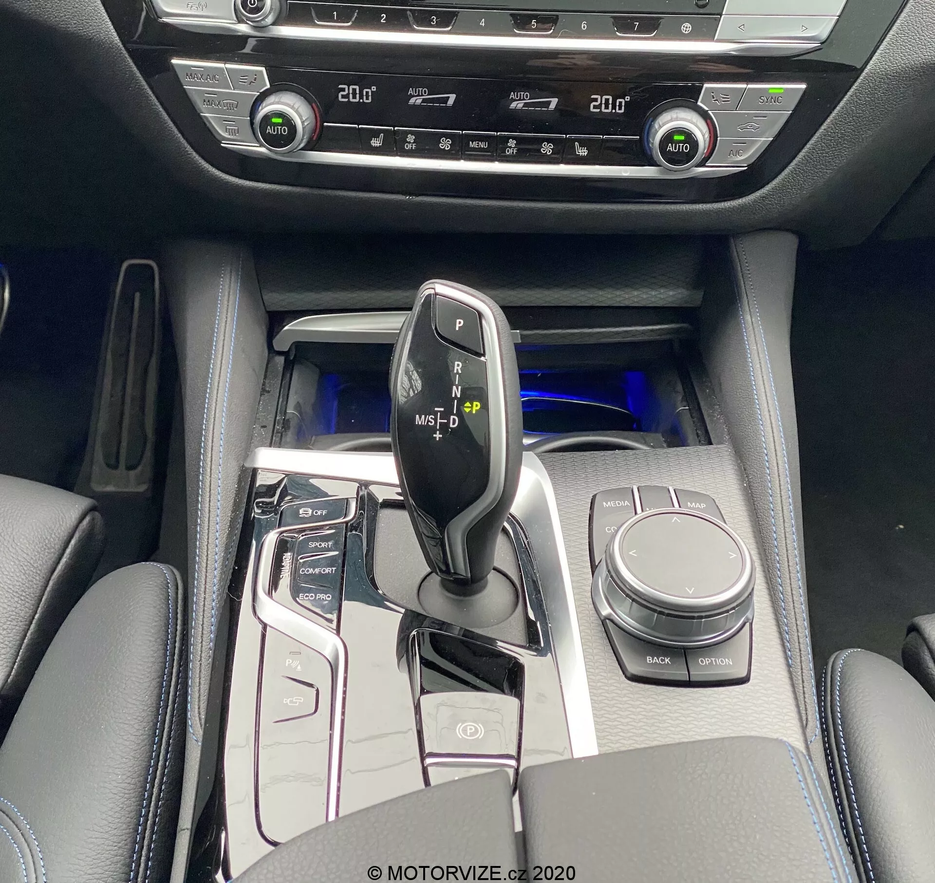 Vue en plongée de la console centrale de la BMW 520i Touring Steptronic 2020, capturant un levier de vitesse noir brillant avec des accents métalliques, entouré de boutons de panneau de contrôle haute brillance pour système start/stop, modes de conduite, capteurs de stationnement et plus. Visible également, le bouton de contrôle iDrive, les porte-gobelets et l'accoudoir rembourré avec rangement. L'éclairage d'ambiance bleu froid rehausse le sentiment de luxe. Les commandes bi-zone de climatisation et de chauffage des sièges indiquent des fonctions de confort avancées. Les matériaux intérieurs comprennent des surfaces similicuir avec accents métalliques et panneaux à haute brillance.