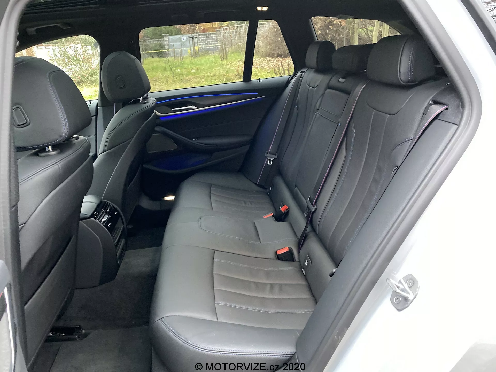 Intérieur de la BMW 520i Touring Steptronic 2020 focalisé sur l'espace des sièges arrière depuis le côté droit. Le point de vue est près de la porte arrière droite du passager, montrant une banquette pour trois avec une sellerie en cuir foncé et des surpiqûres décoratives. Les caractéristiques incluent trois appuie-têtes, trois ceintures de sécurité, un panneau de porte avec une grille de haut-parleur, une poignée et des éléments de contrôle, une bande d'éclairage d'ambiance bleue et un filet de rangement à l'arrière du siège passager avant. L'intérieur bien entretenu ne présente ni effets personnels visibles ni usure significative. La vue partielle à l'extérieur par la fenêtre arrière inclut une clôture.