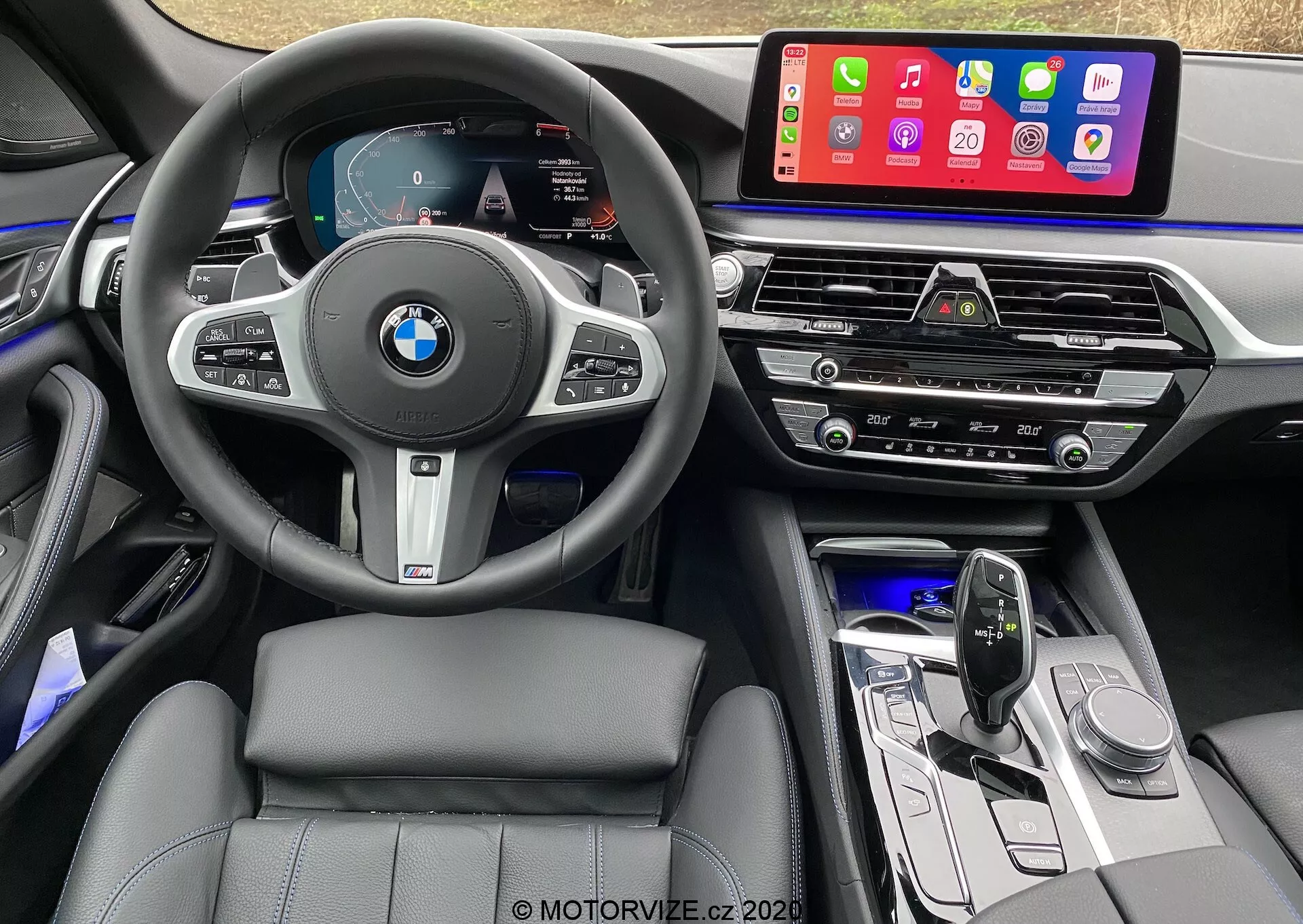 Vue intérieure depuis le siège du conducteur d'une BMW 520i Touring Steptronic 2020, comprenant un volant multifonction en cuir avec le logo BMW et des boutons de contrôle pour le régulateur de vitesse, l'audio et la téléphonie. Le tableau de bord numérique affiche le tachymètre, le compteur de vitesse et une carte de navigation. La console centrale est équipée d'un grand système d'infodivertissement à écran tactile affichant des applications mobiles comme Apple CarPlay. En dessous se trouvent les bouches d'aération, le panneau de contrôle de la climatisation, les commandes audio, le levier de vitesses au design élégant et les boutons de fonction. L'intérieur présente des sièges cousus en cuir, un éclairage d'ambiance et des matériaux de haute qualité avec des accents métalliques.