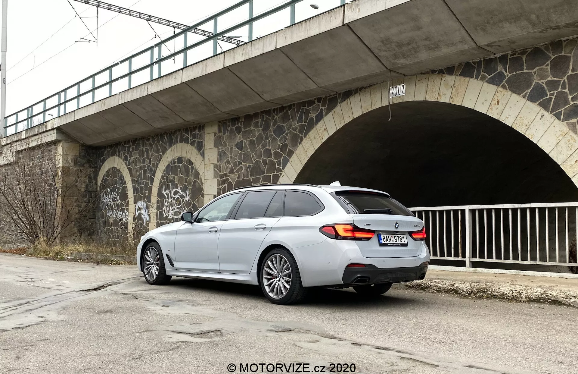 L'image montre une voiture break moderne, de couleur argentée, BMW Série 5 Touring (520i Touring Steptronic) stationnée sur une surface asphaltée, offrant une vue arrière trois quarts. Le véhicule est positionné sur le côté droit du cadre, presque parallèle au bord inférieur de l'image. L'arrière de la voiture, faisant face à la caméra, montre les feux arrière, le hayon, le pare-chocs arrière et la majeure partie du côté droit, y compris la porte arrière côté passager, la roue arrière droite et une partie de la porte et de la roue avant droite. Les éléments de design distinctifs incluent les feux arrière, le système d'échappement double, les jantes en alliage à cinq branches, la grande lunette arrière et le spoiler de toit. L'environnement urbain comprend un pont en hauteur et une entrée de tunnel avec des graffitis et des éléments de maçonnerie, sous un ciel couvert.