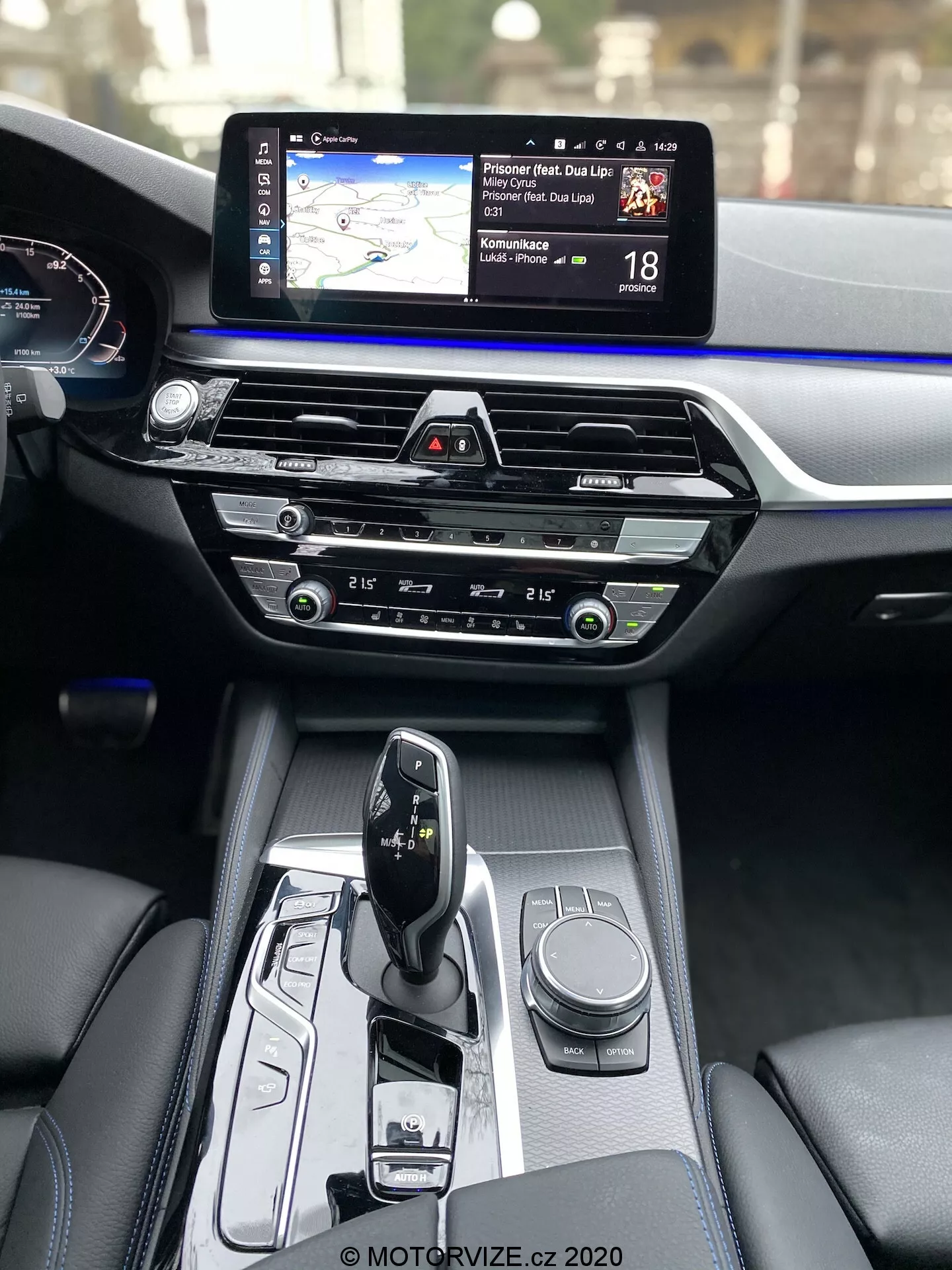 Vue intérieure de la BMW Série 5 2020 (BMW 520i Touring Steptronic), montrant la console centrale et le tableau de bord depuis la perspective du passager avant. La console centrale comprend un sélecteur de vitesse automatique en mode 'park', avec divers contrôles incluant un cadran rotatif pour le système d'infodivertissement et des boutons pour les modes de conduite ou les réglages du véhicule. La section centrale du tableau de bord inclut des commandes de climatisation avec des cadrans et des boutons intuitifs, au-dessus desquels se trouve une fente pour CD. La partie supérieure du tableau de bord présente un large écran tactile d'infodivertissement avec une carte de navigation et des informations sur la limite de vitesse. Partiellement visible se trouve le tableau de bord numérique montrant le compteur de vitesse. Les matériaux intérieurs mettent en valeur des finitions de luxe, contrastant le cuir sombre avec une bordure bleue tout le long du tableau de bord.