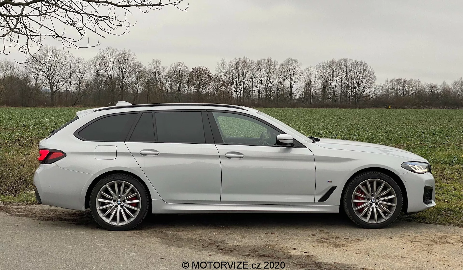 Profil latéral d'une BMW 520i Touring Steptronic break moderne capturé en pleine lumière du jour, mettant en avant des lignes de design élégantes, cinq portes dont le hayon arrière, une longue ligne de toit avec un spoiler, de grandes jantes en alliage à plusieurs rayons avec des étriers de frein visibles, des poignées de porte de la couleur de la carrosserie, des clignotants intégrés aux rétroviseurs latéraux, des vitres légèrement teintées et une finition de peinture mate ou non métallique. La voiture est positionnée sur l'accotement en gravier ou en terre à côté d'une route pavée dans un environnement rural, avec de l'herbe et des arbres en arrière-plan, dans des conditions météorologiques clémentes.