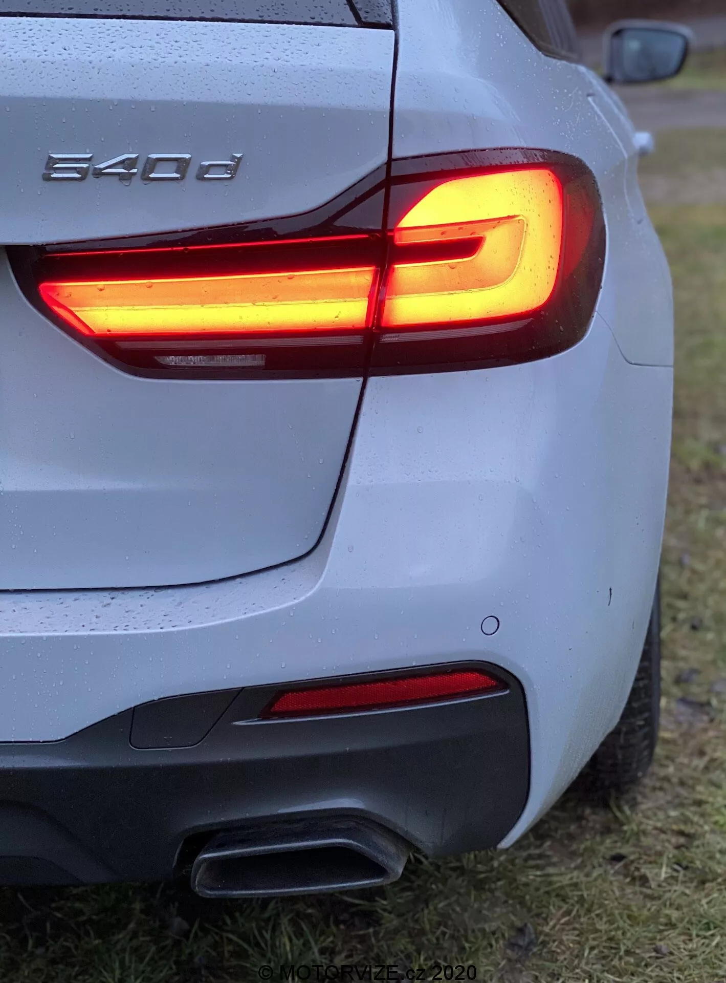 Vue en angle arrière trois quarts rapprochée d'une BMW 540i, montrant le feu arrière en L à LED, les doubles sorties d'échappement, la carrosserie argentée/grise avec des gouttes d'eau, le pare-chocs arrière sculpté, le badge partiel de BMW, stationnée sur une zone herbeuse.