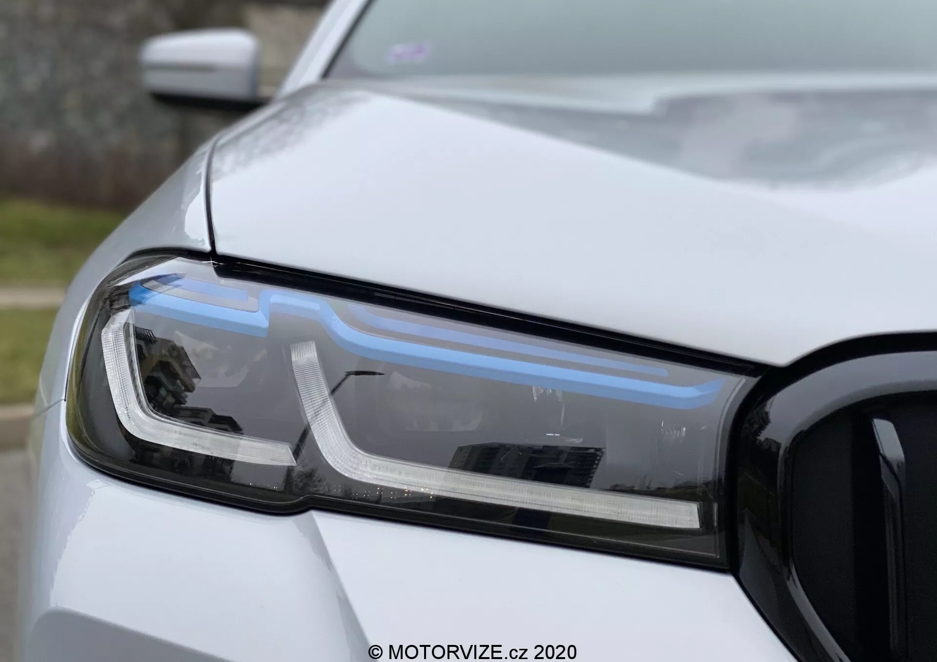 Vue rapprochée de l'ensemble du phare côté conducteur d'une BMW 520i Touring Steptronic, mettant en avant les feux de jour à LED, le phare à projecteur et l'indicateur de direction. La photographie montre la partie avant gauche de la voiture, y compris une partie du capot, du pare-chocs avant et de l'aile, avec la calandre visible sur la droite. La prise de vue présente une faible profondeur de champ mettant en valeur la technologie et le design du phare dans une lumière naturelle diffuse avec une finition de peinture blanche non métallique.