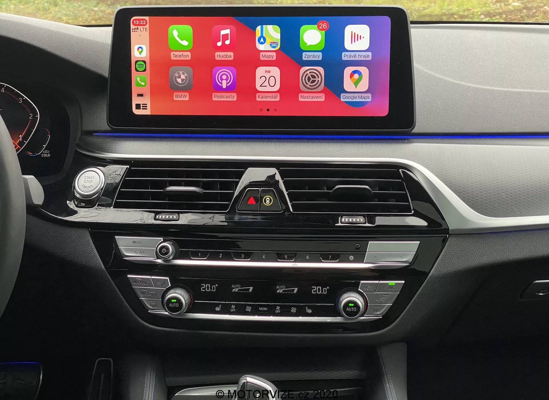 Vue intérieure d'une BMW 5er-Reihe 2020 (BMW 520i Touring Steptronic) depuis la perspective du siège conducteur, se concentrant sur le tableau de bord central. La caractéristique la plus notable est un grand écran tactile d'infodivertissement horizontal avec une interface colorée supportant l'intégration de smartphones comme Apple CarPlay et Android Auto. Sous l'écran, des évents d'air avec des accents chromés et un bouton de feux de détresse sont visibles. Plus bas, on trouve les contrôles de climatisation bi-zone avec des affichages numériques pour les réglages de température, ainsi que des boutons et des cadrans pour ajuster la température de l'air, la vitesse du ventilateur et la distribution de l'air. Le tableau de bord et la console centrale comportent des accents plastiques et métalliques souples au toucher, augmentant l'attrait esthétique.